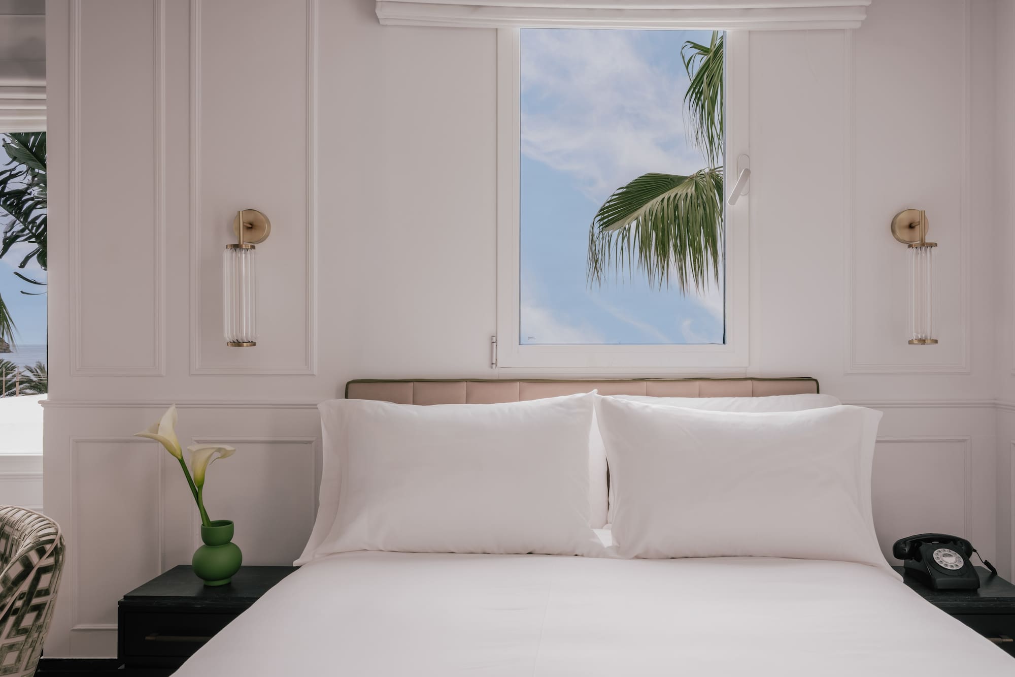 Hotel bedroom with white bed, palm tree outside window, bedside phone.