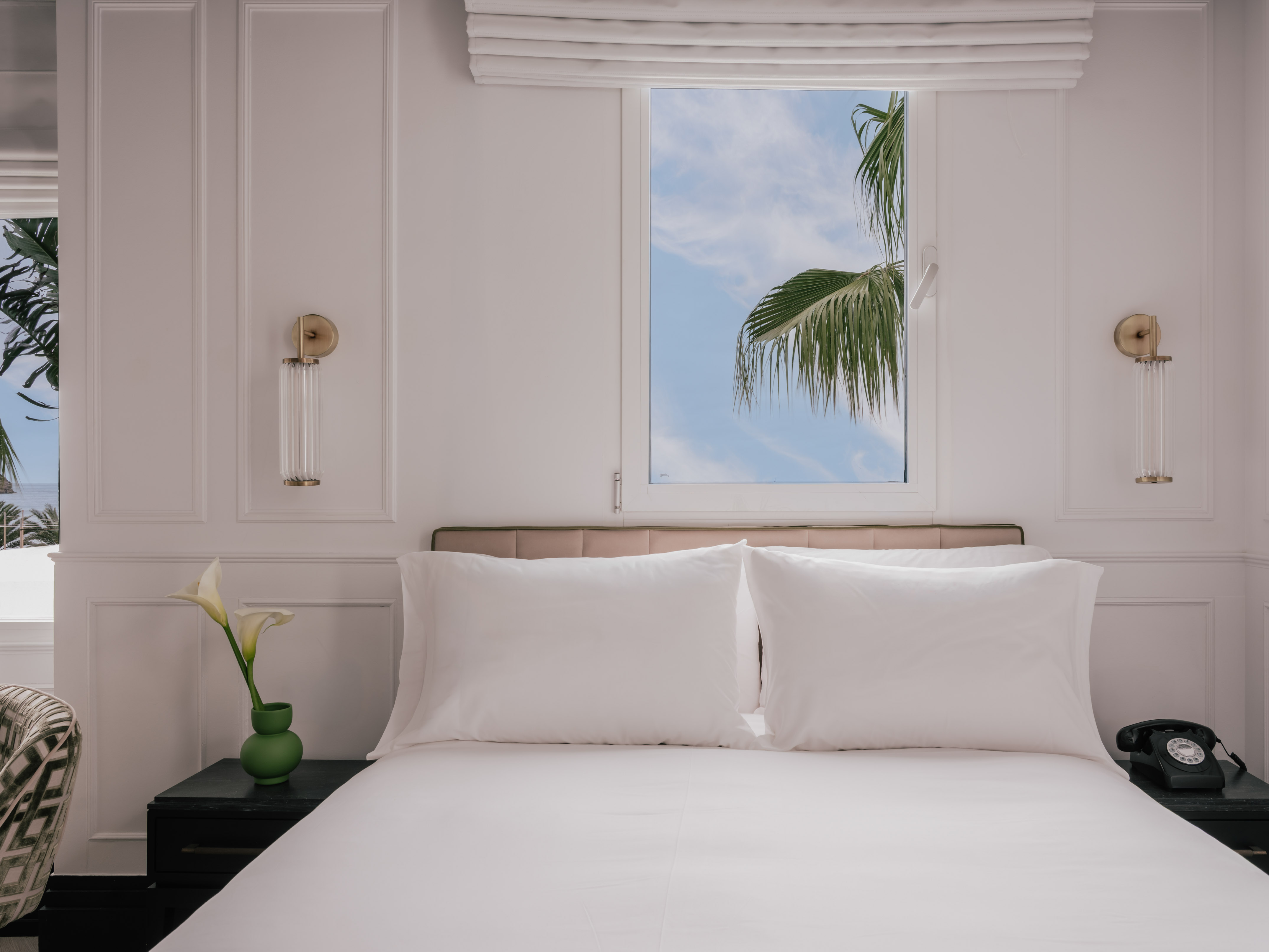 Hotel bedroom with white bed, palm tree outside window, bedside phone.