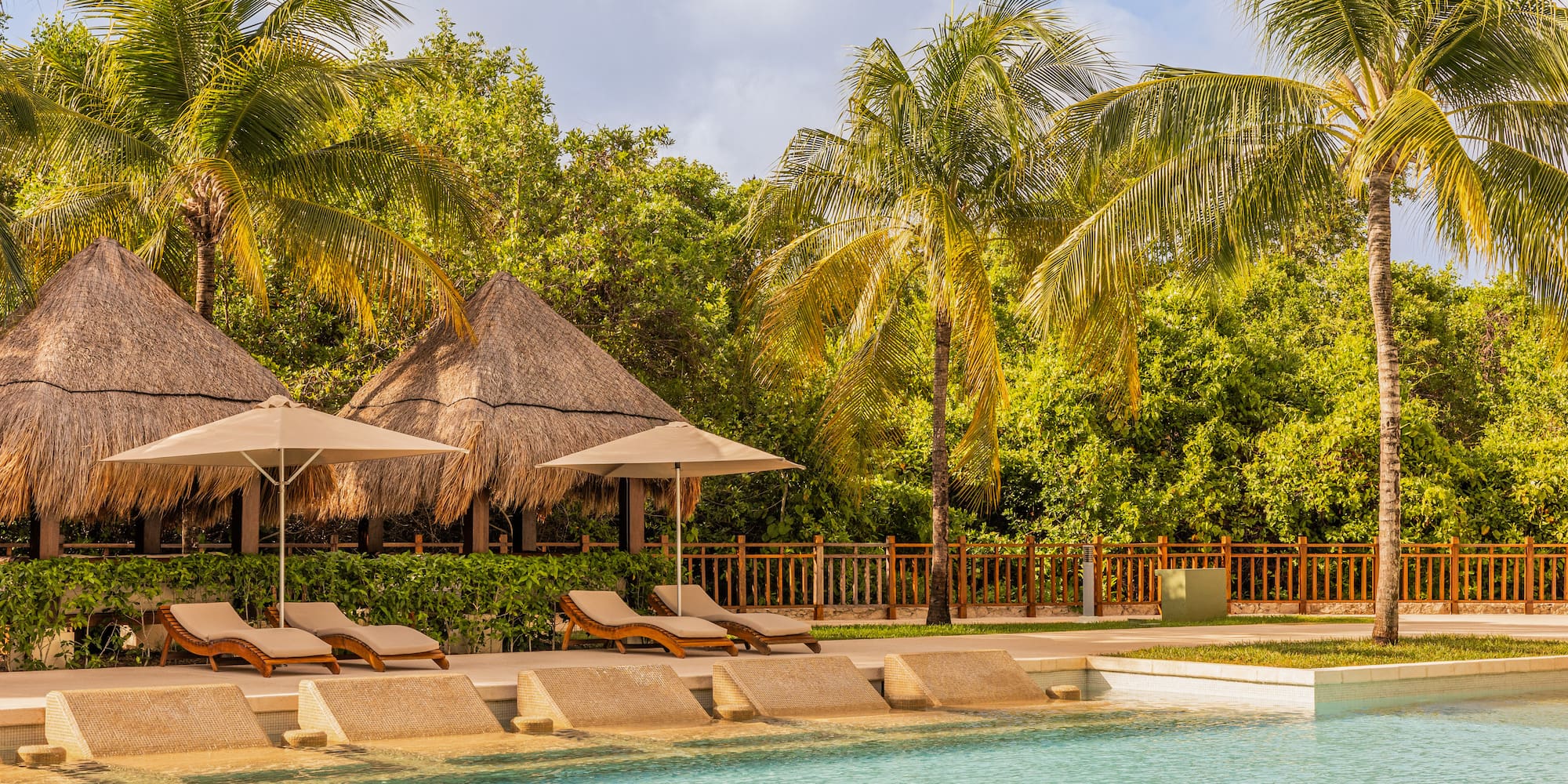 Tropical swimming pool with lounge chairs, thatched palapas, and palm trees.