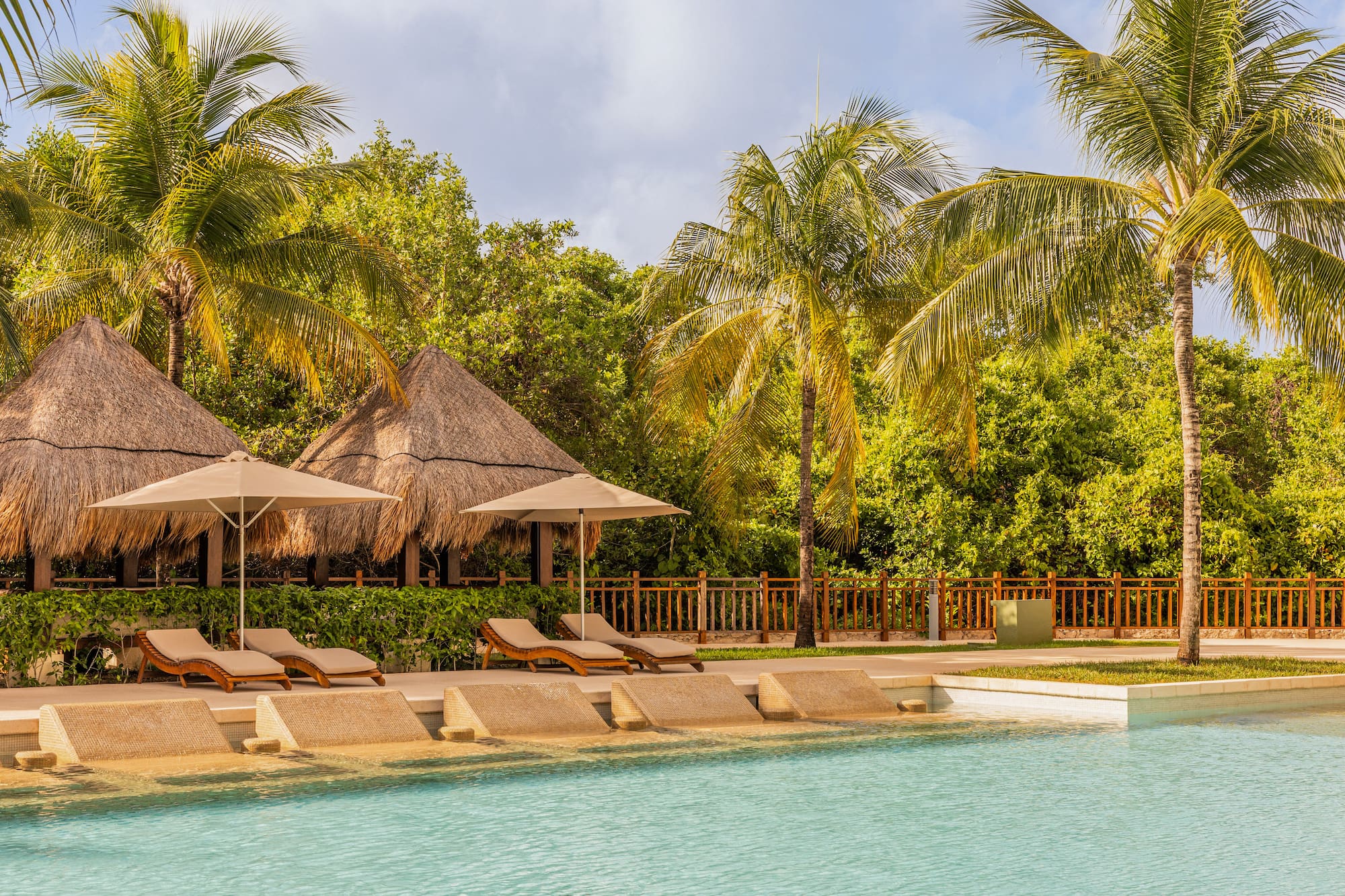 Tropical swimming pool with lounge chairs, thatched palapas, and palm trees.