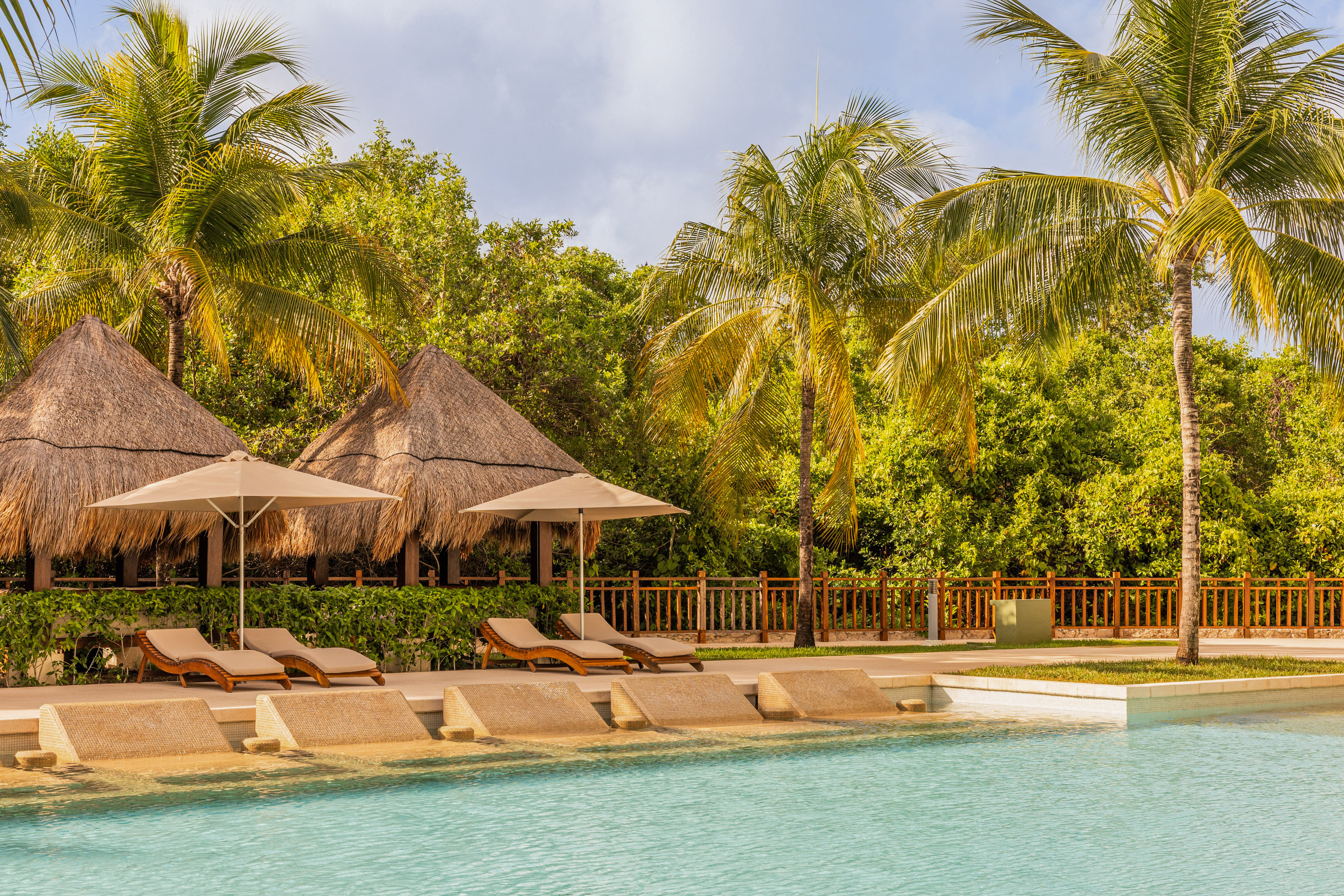 Tropical swimming pool with lounge chairs, thatched palapas, and palm trees.