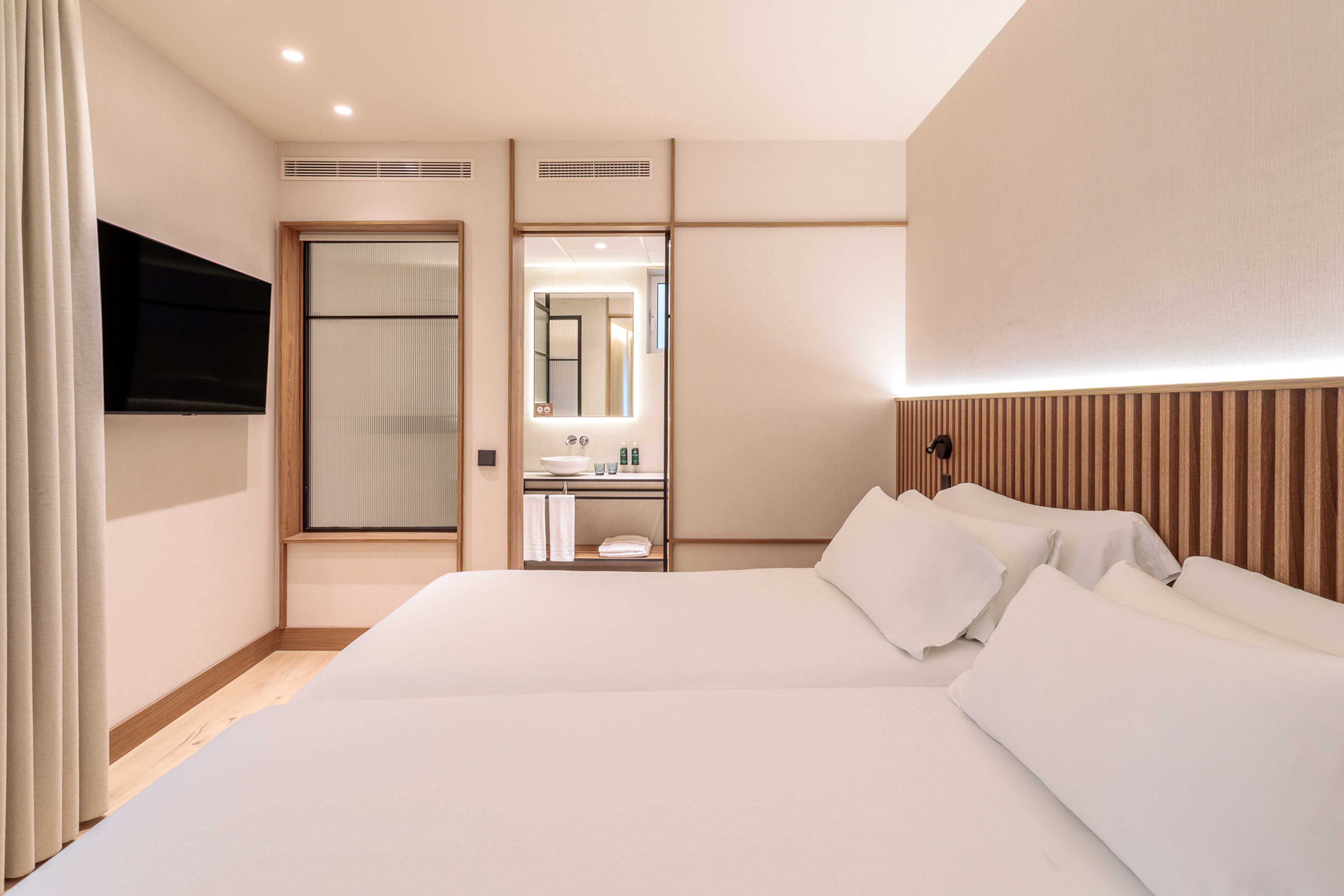 Modern hotel bedroom with twin beds, wooden headboard, TV, and open vanity.