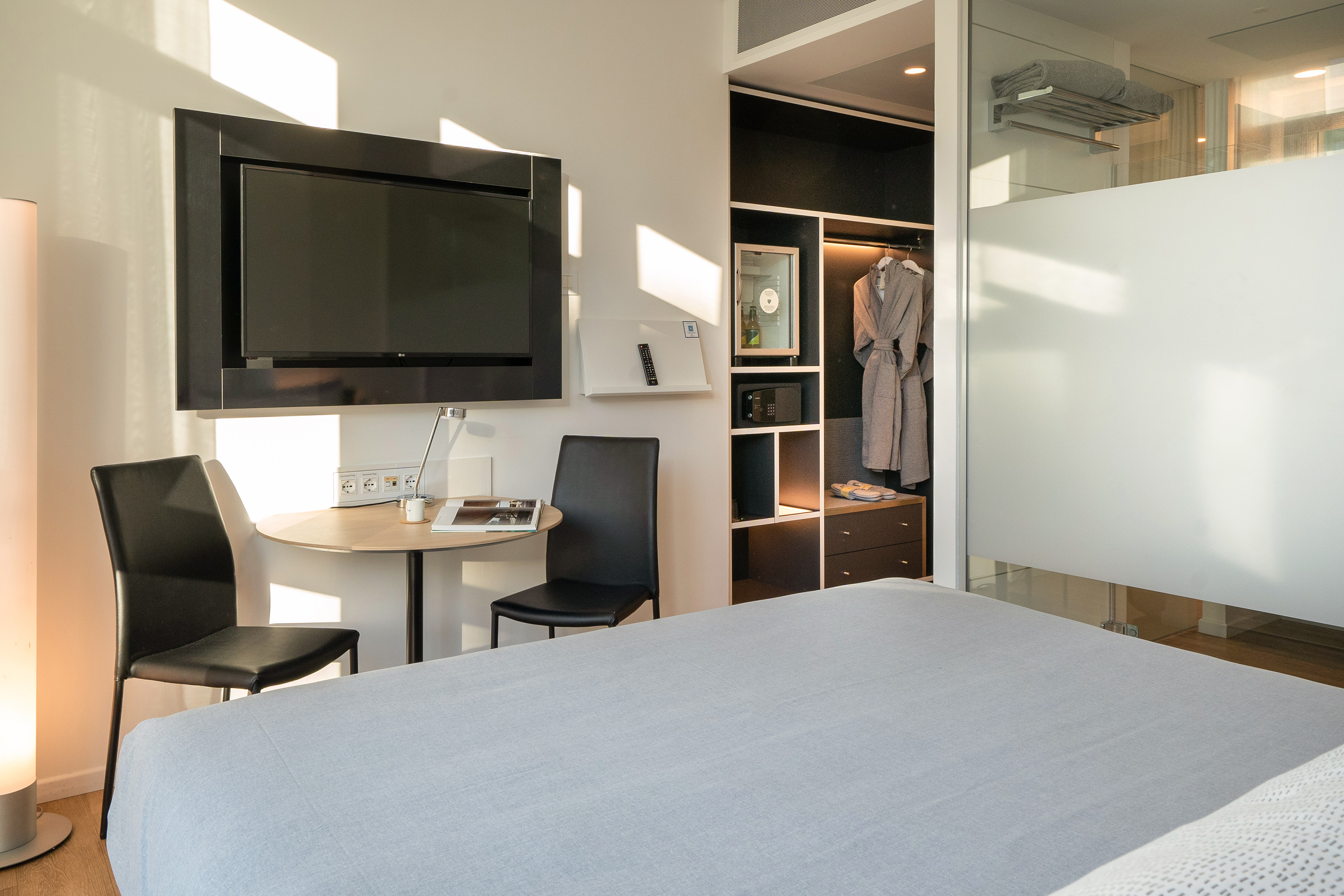 Modern hotel room with bed, wall TV, desk, and open closet featuring robes.