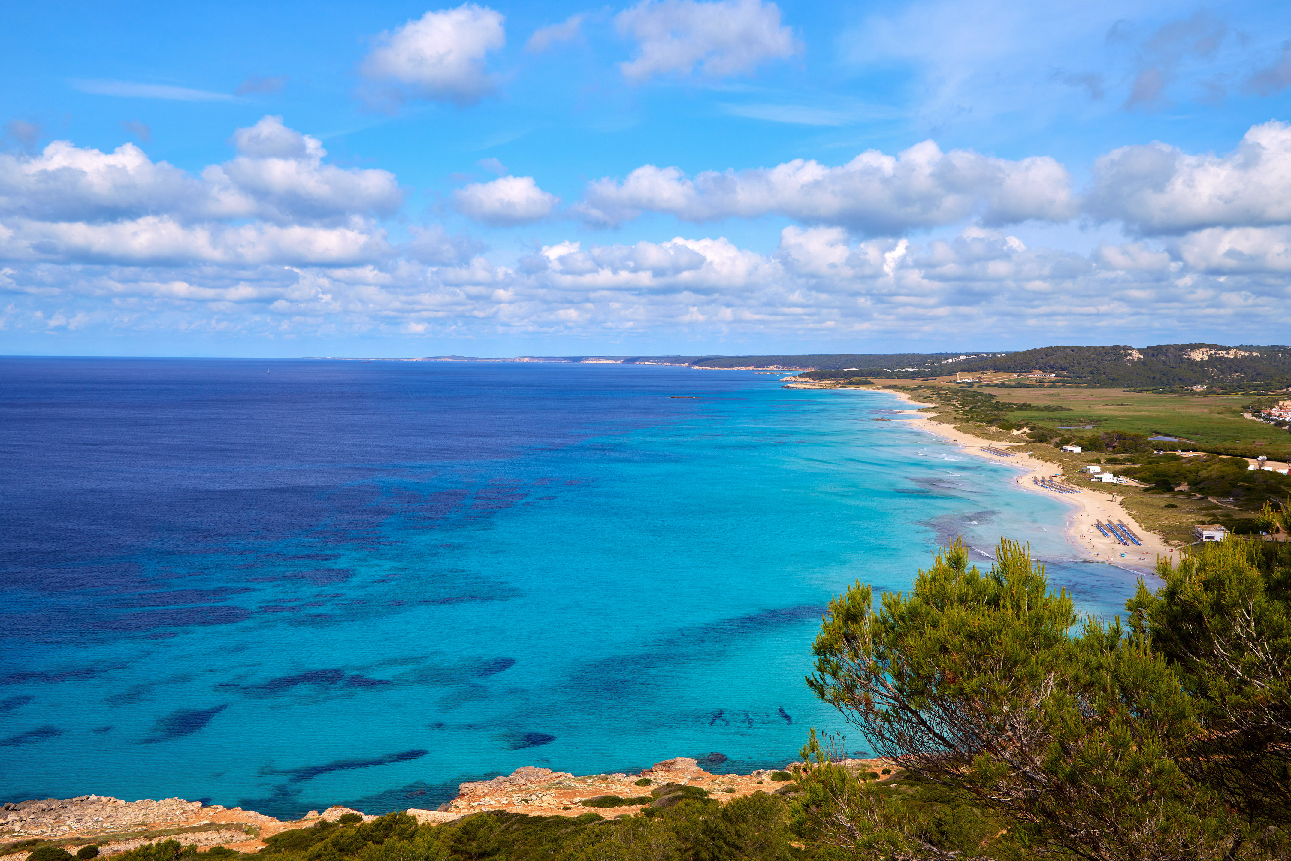 Turquoise sea coastline with sandy beach, green vegetation, and blue sky.