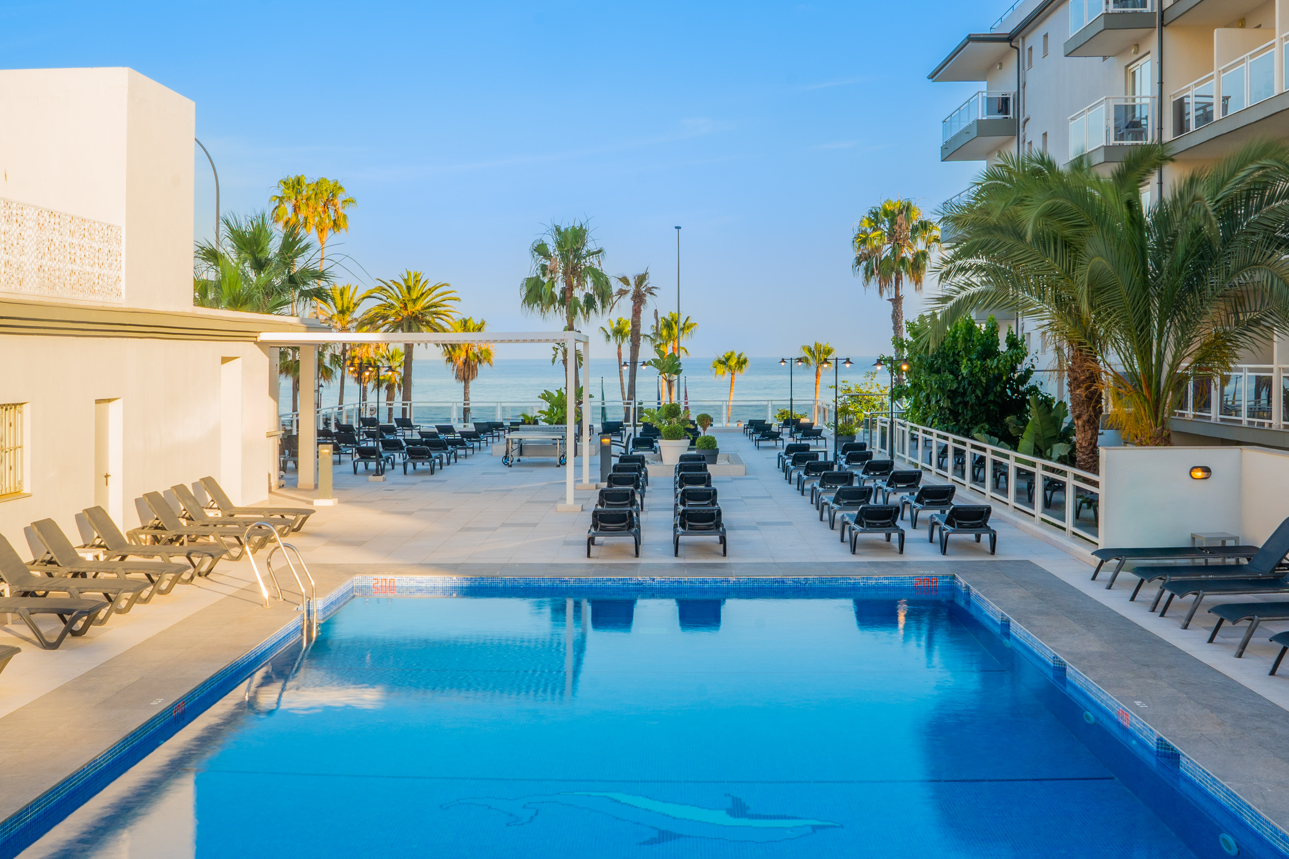 Tropical resort pool area with sea view, palm trees, and sunbeds.