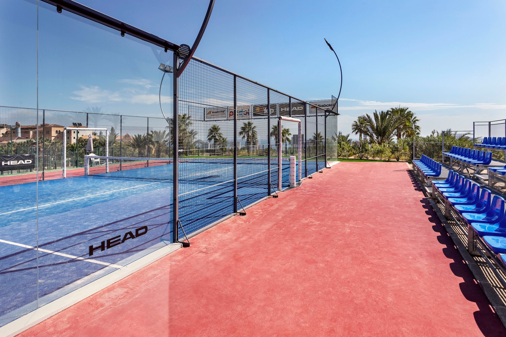 Sunny outdoor padel court with blue surface, glass walls, red spectator seats.