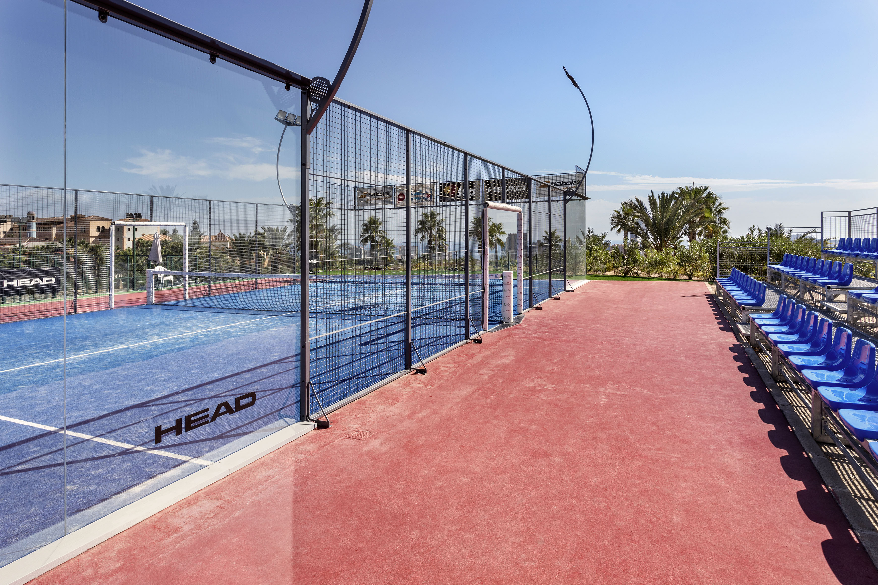 Sunny outdoor padel court with blue surface, glass walls, red spectator seats.