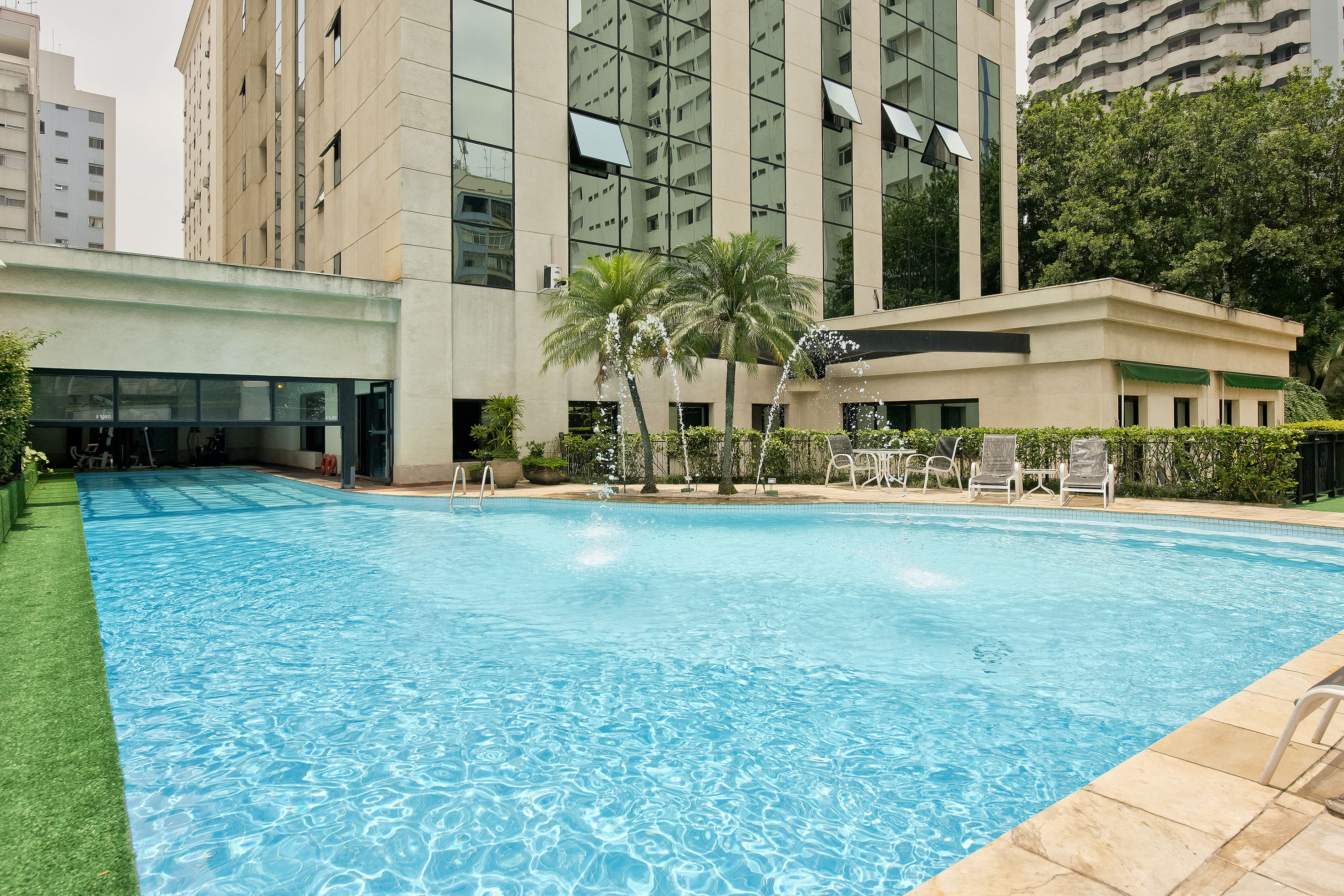 Outdoor hotel swimming pool with palm trees, fountains, and modern building.