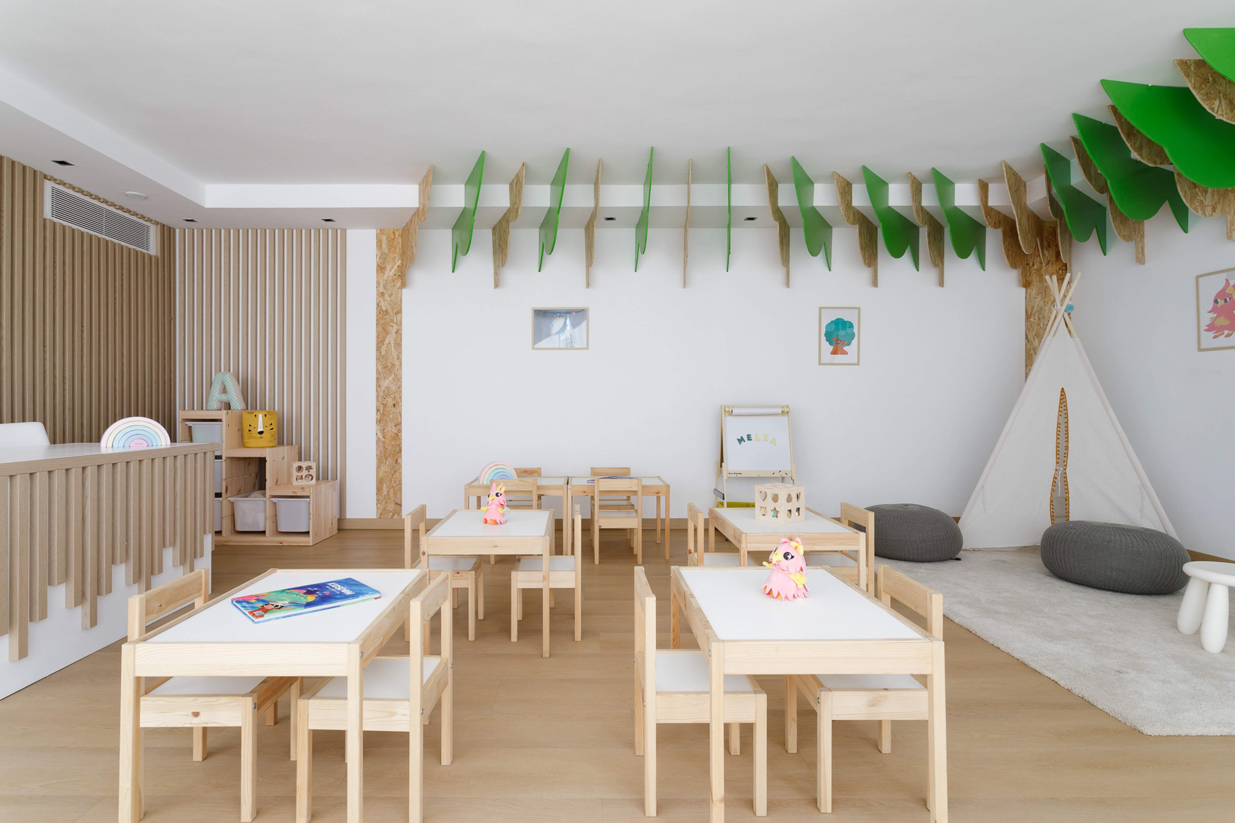 Bright preschool classroom with wooden tables, leaf ceiling decor, and teepee.