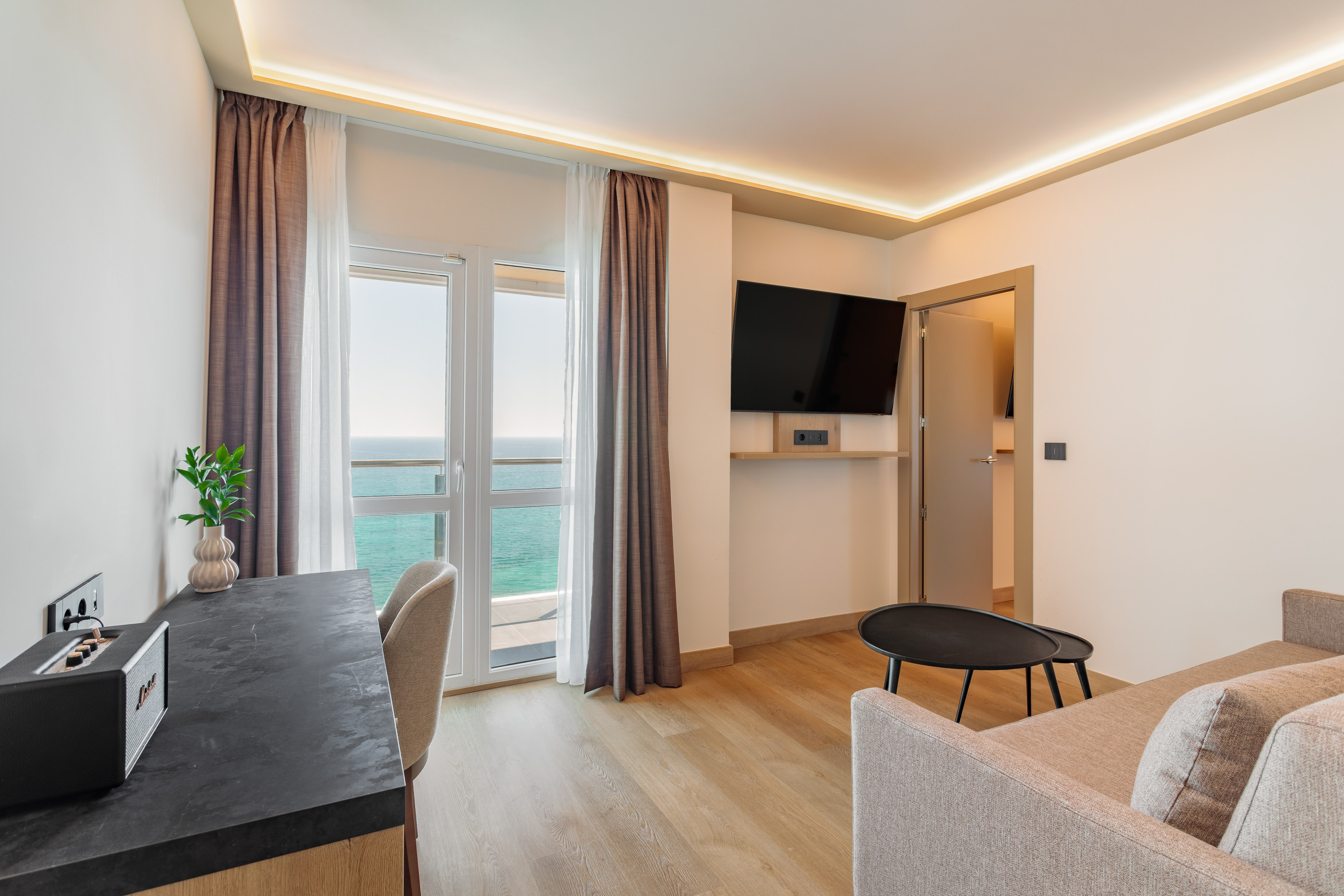 Modern hotel suite with ocean view balcony, desk, TV, and sofa.