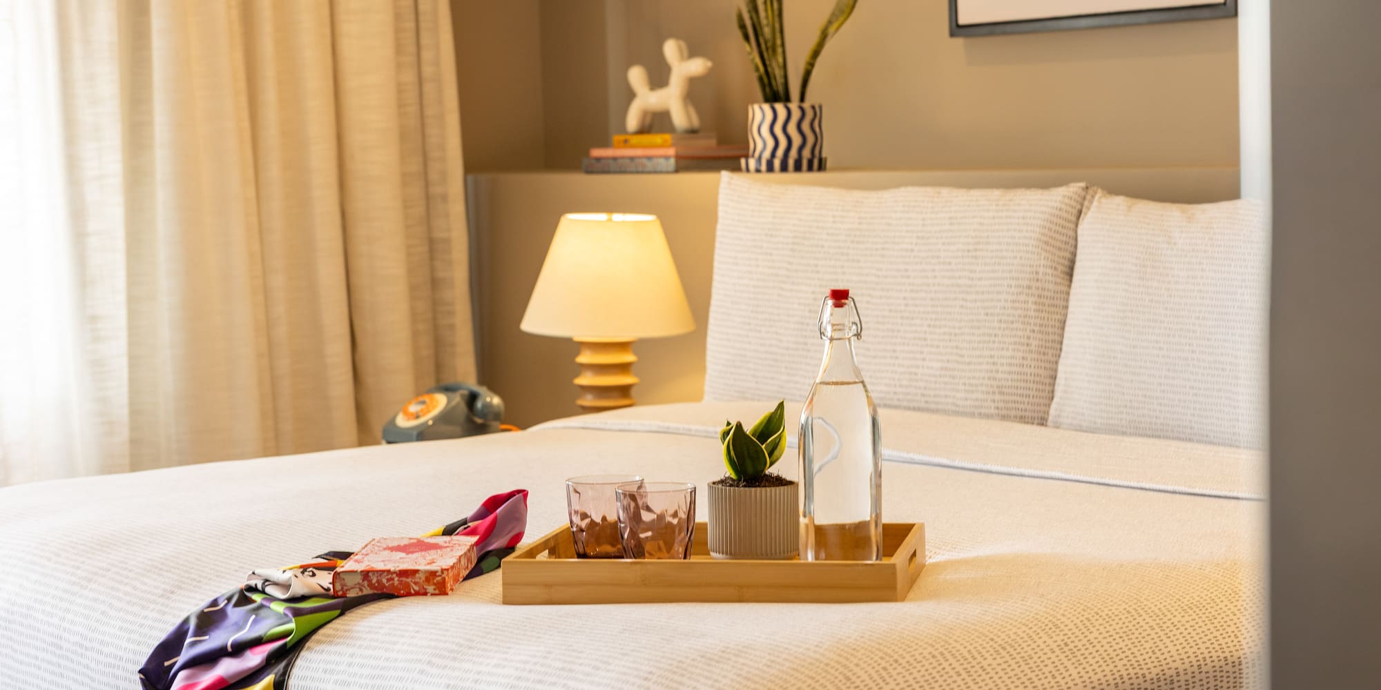 Stylish hotel room bed with water, plant on tray, colorful scarf, bedside lamp.