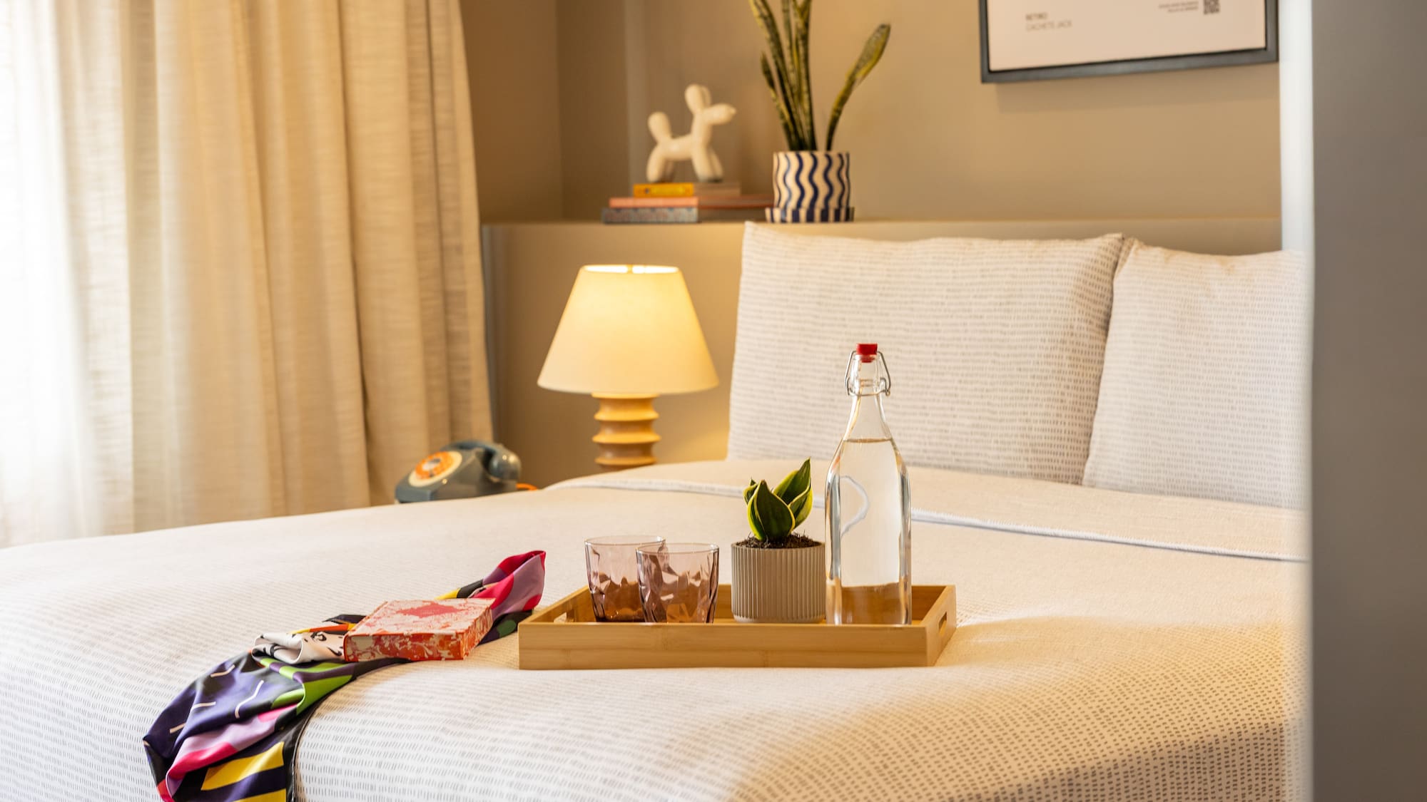 Stylish hotel room bed with water, plant on tray, colorful scarf, bedside lamp.