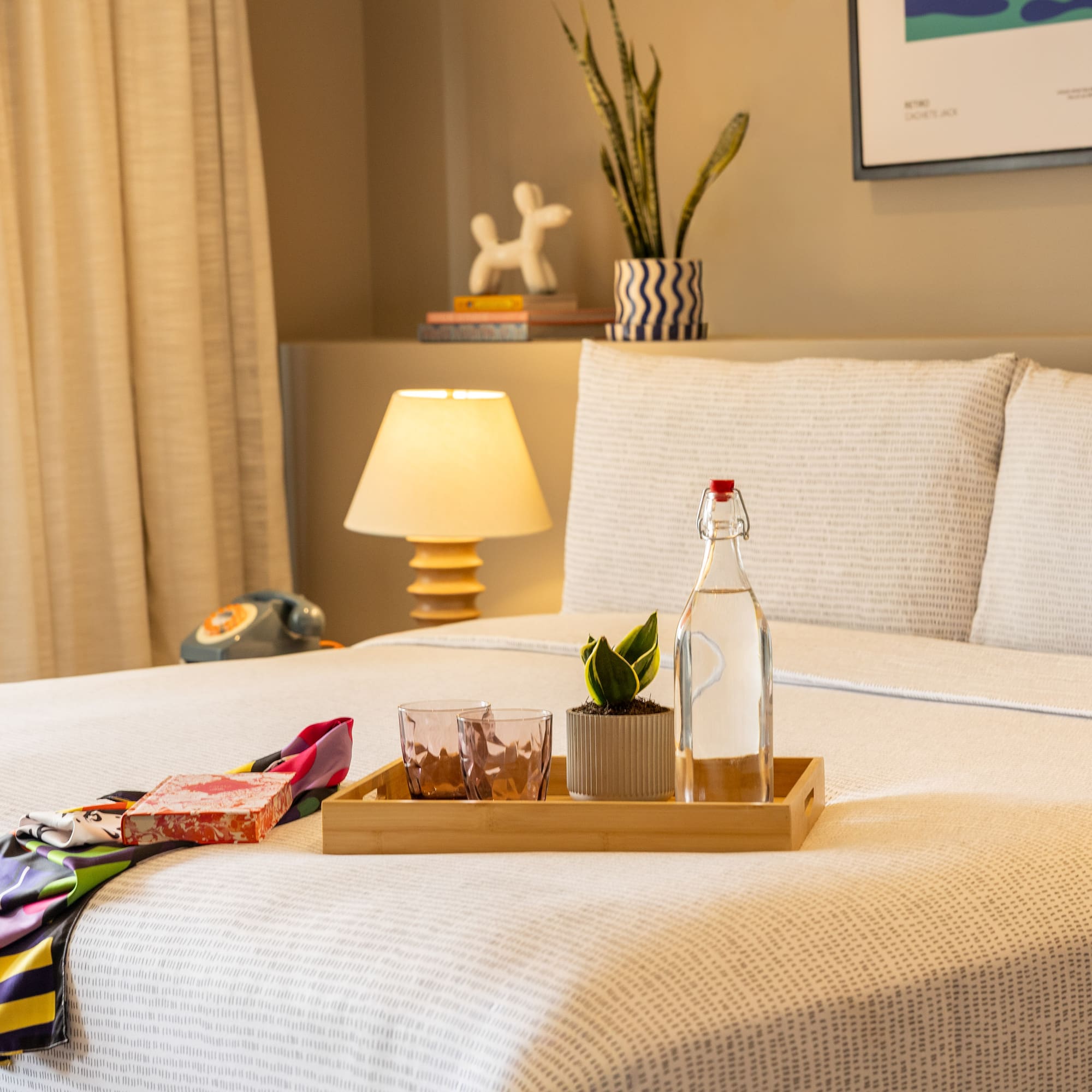 Stylish hotel room bed with water, plant on tray, colorful scarf, bedside lamp.