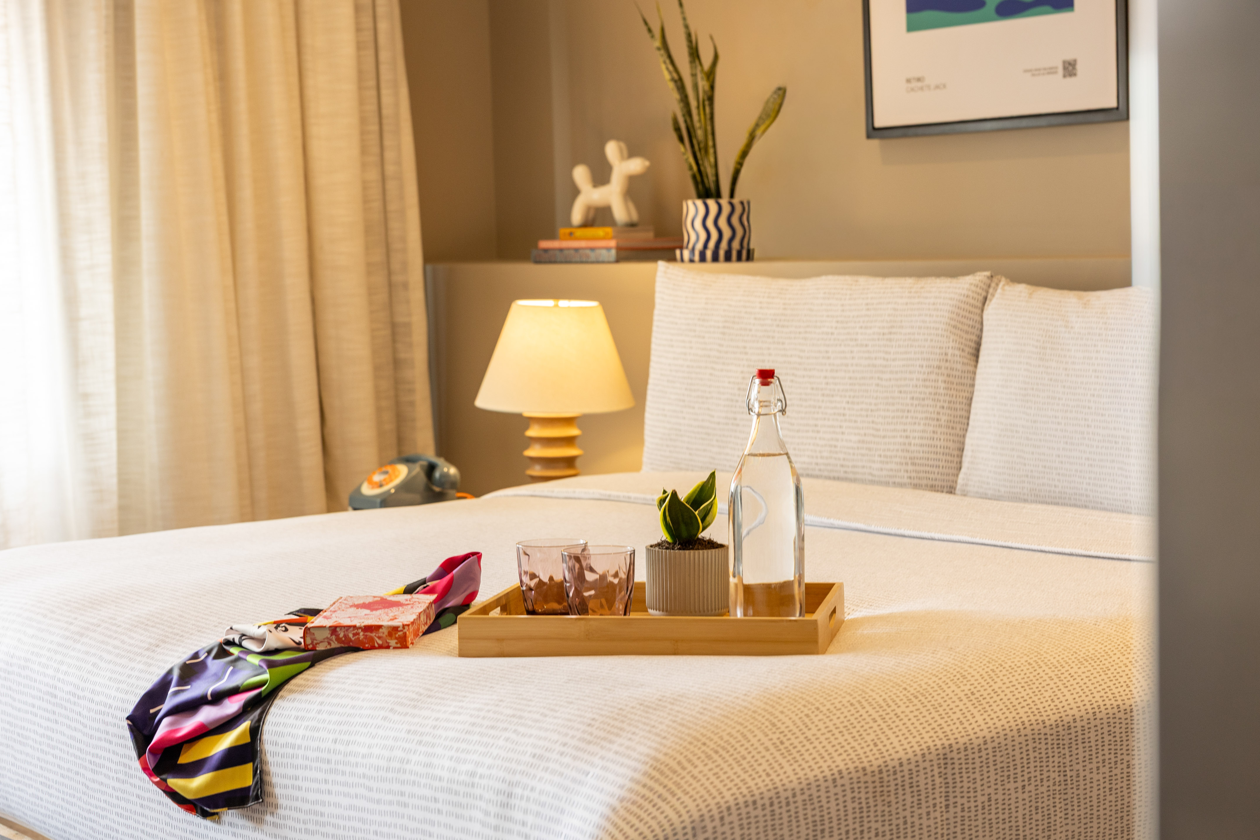 Stylish hotel room bed with water, plant on tray, colorful scarf, bedside lamp.