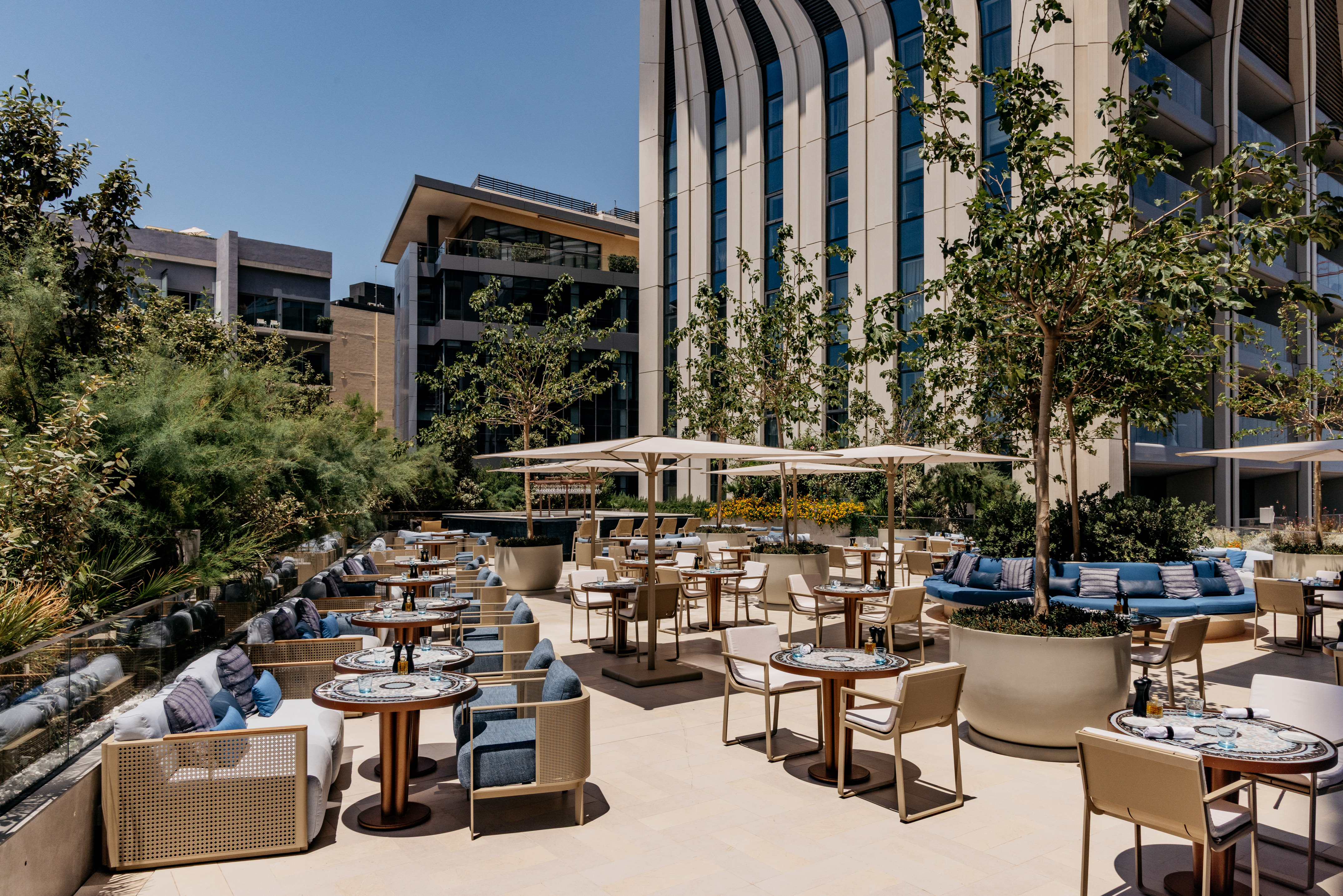 Sunny outdoor dining terrace with modern tables, blue seating, umbrellas, and trees.
