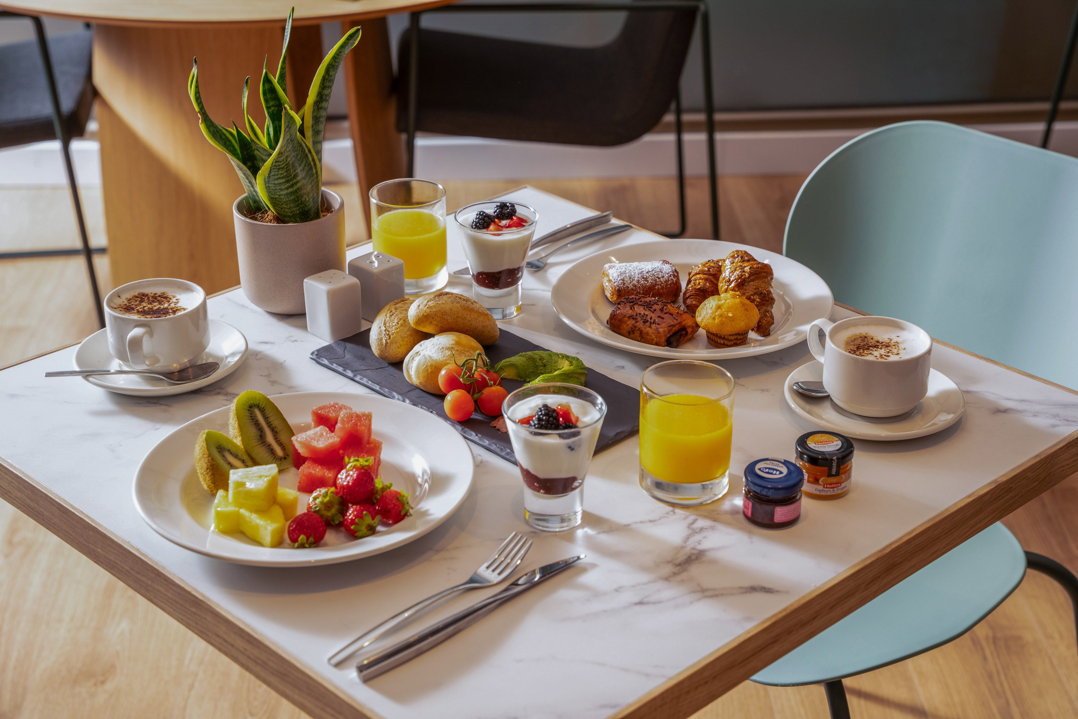 Desayuno variado con café, zumo, bollería, fruta fresca y yogur en mesa.