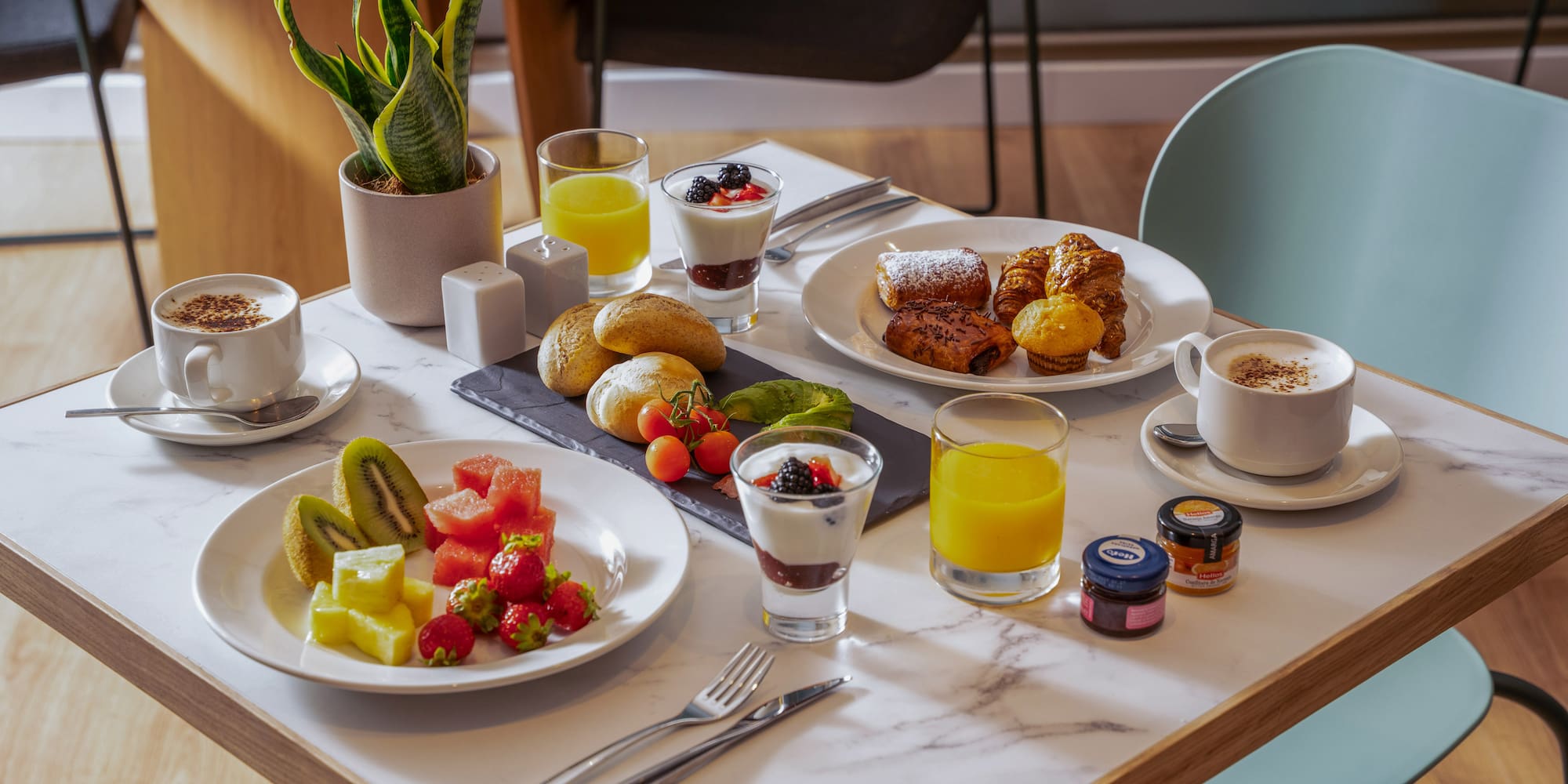 Desayuno variado con café, zumo, bollería, fruta fresca y yogur en mesa.
