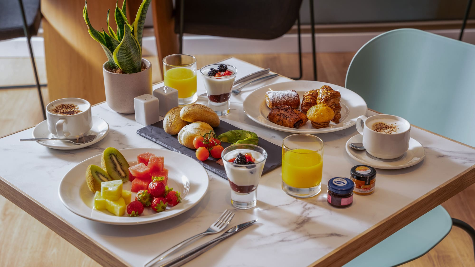 Desayuno variado con café, zumo, bollería, fruta fresca y yogur en mesa.