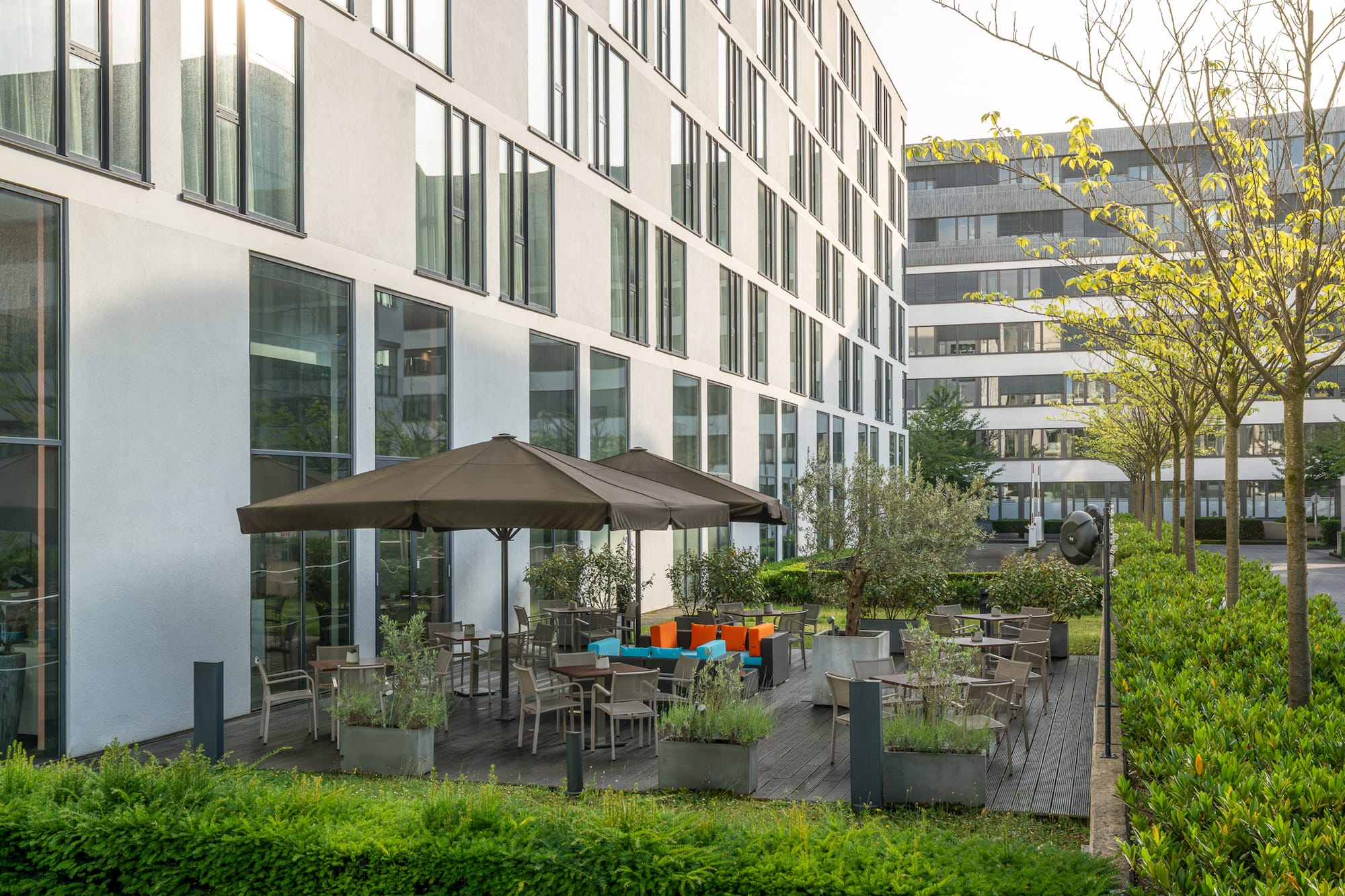 Modern white building with outdoor terrace, shaded seating, and lush green garden.