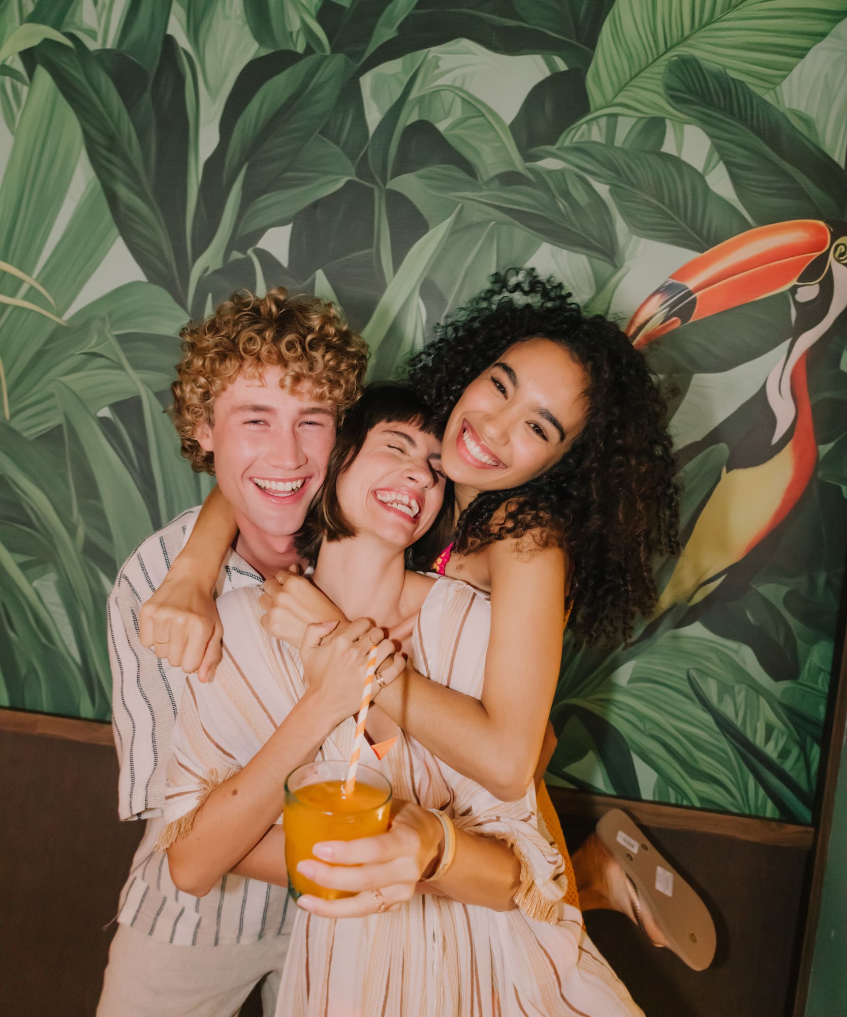 Smiling friends hug, tropical leaf wallpaper with toucan, orange juice.