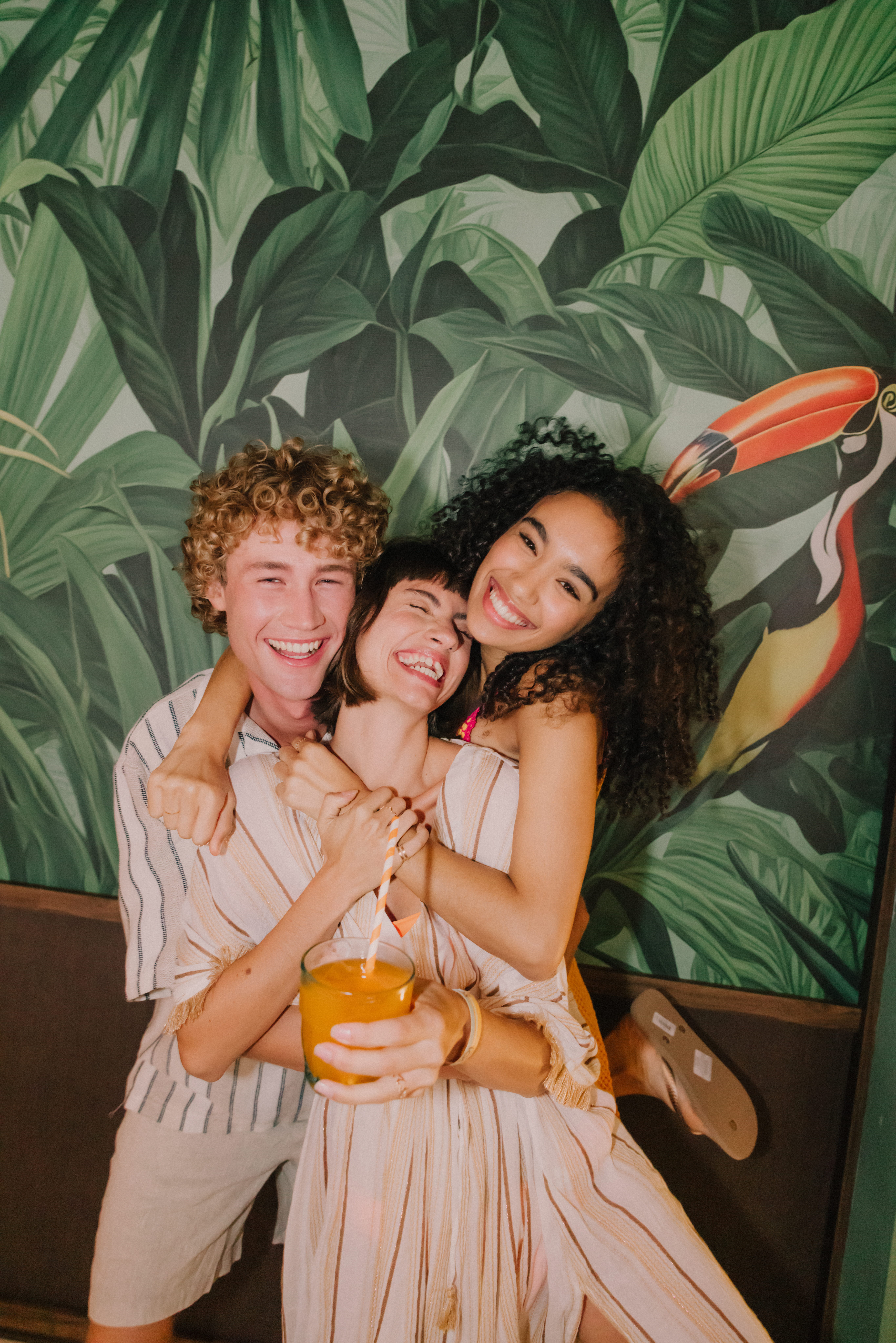 Smiling friends hug, tropical leaf wallpaper with toucan, orange juice.