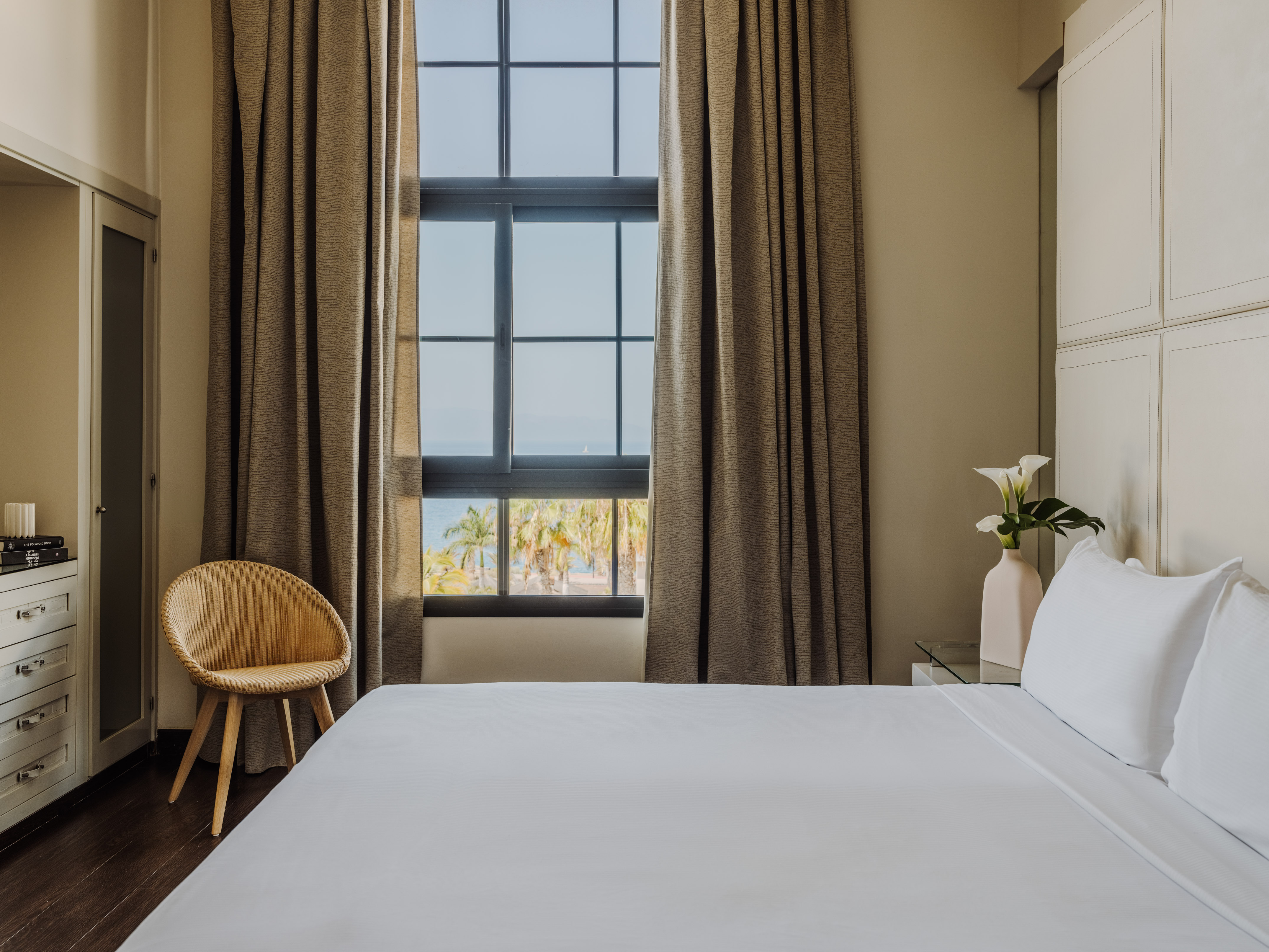 Hotel bedroom with ocean view, white bed, brown curtains, and visible palm trees.