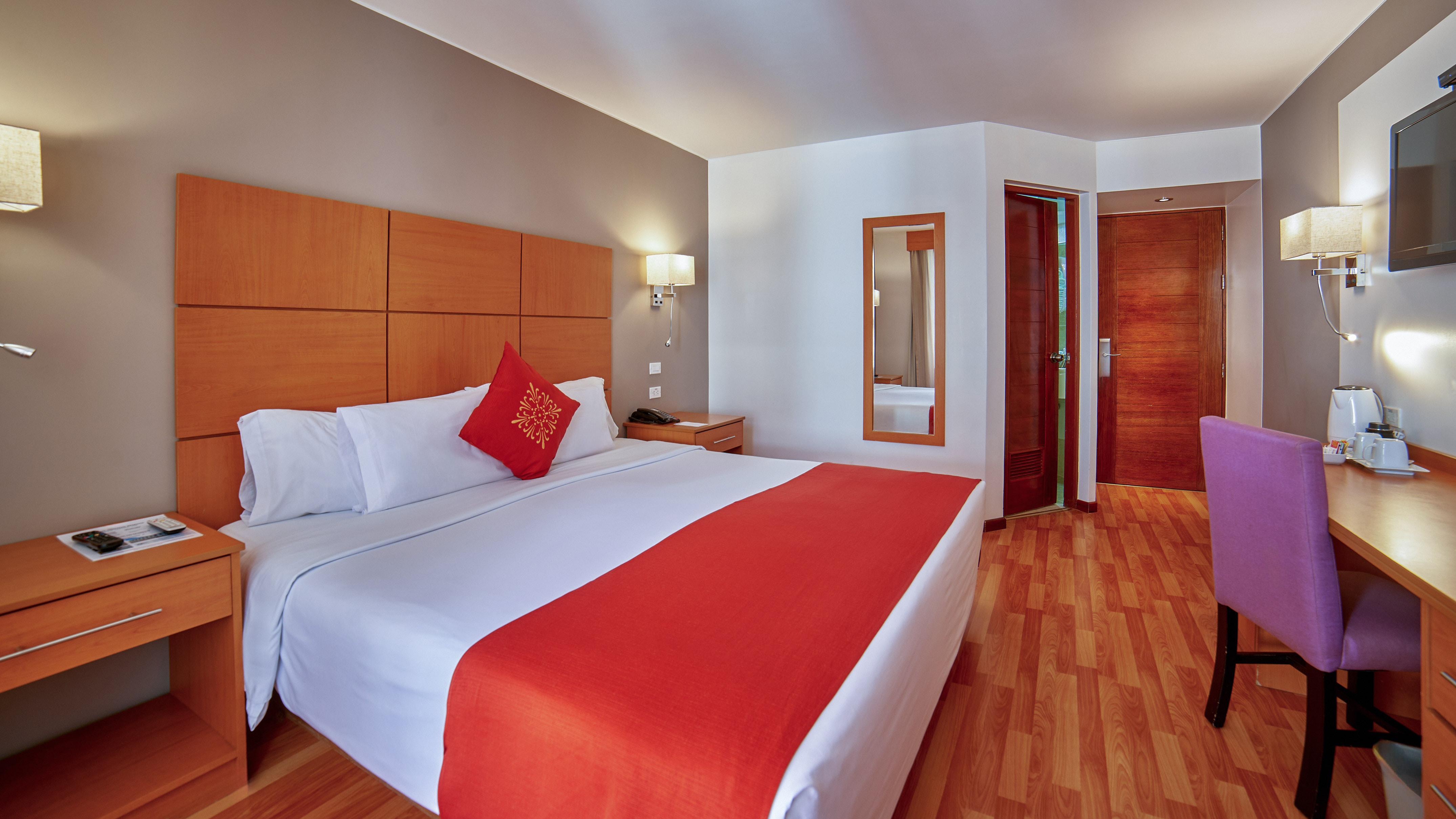 Modern hotel room with king bed, red accents, wooden floor, desk, and TV.