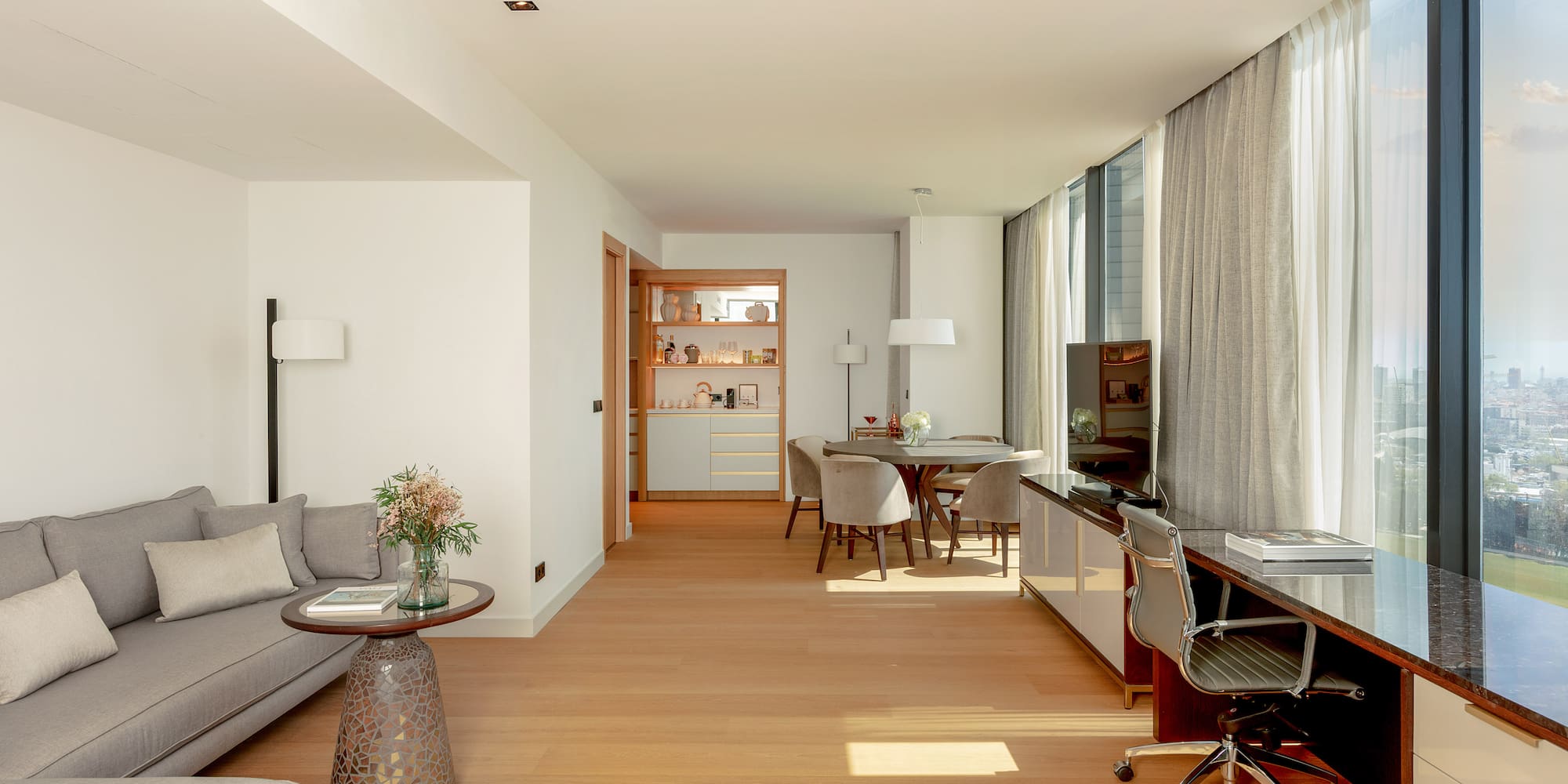 Modern living suite with sofa, dining, desk, hardwood floor, and city view windows.