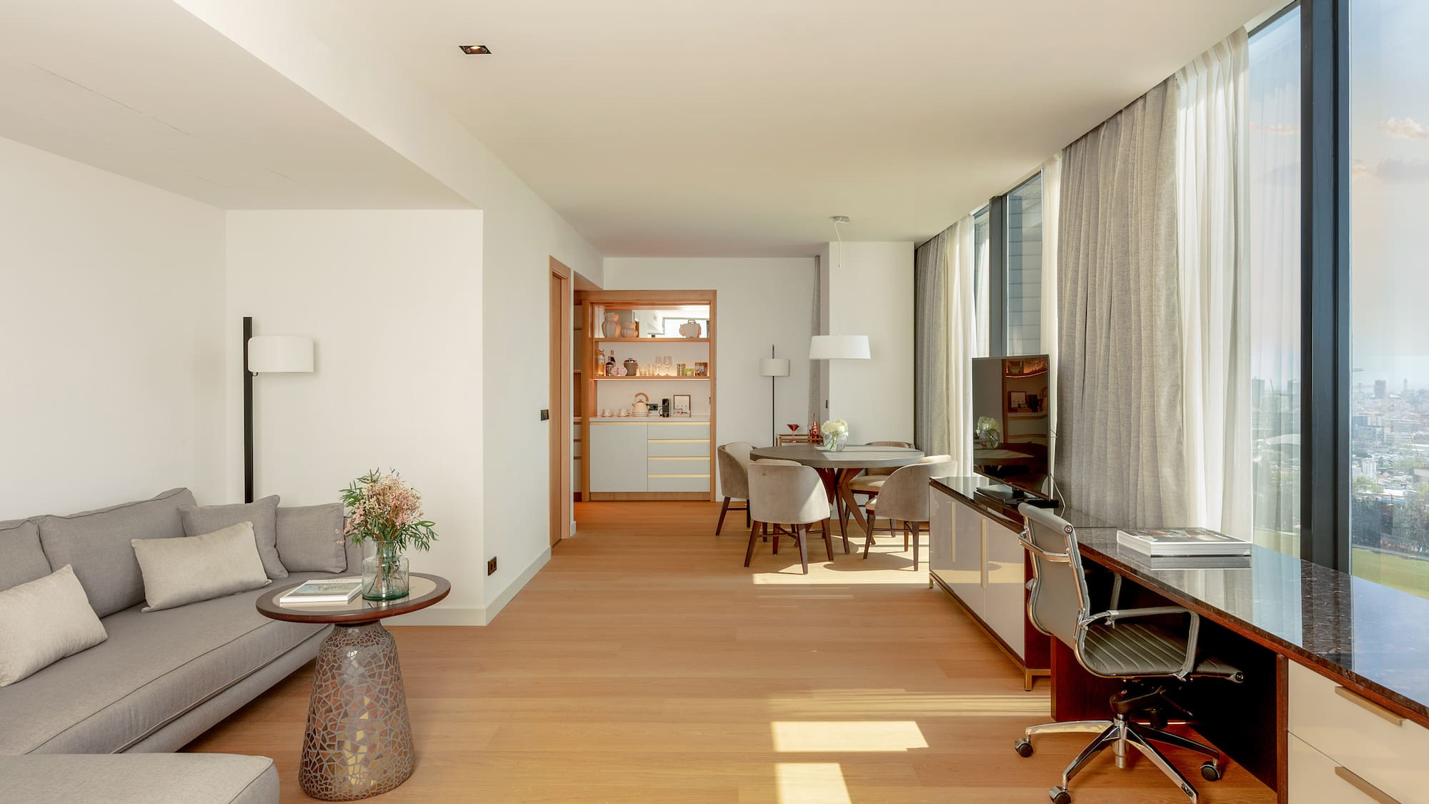 Modern living suite with sofa, dining, desk, hardwood floor, and city view windows.