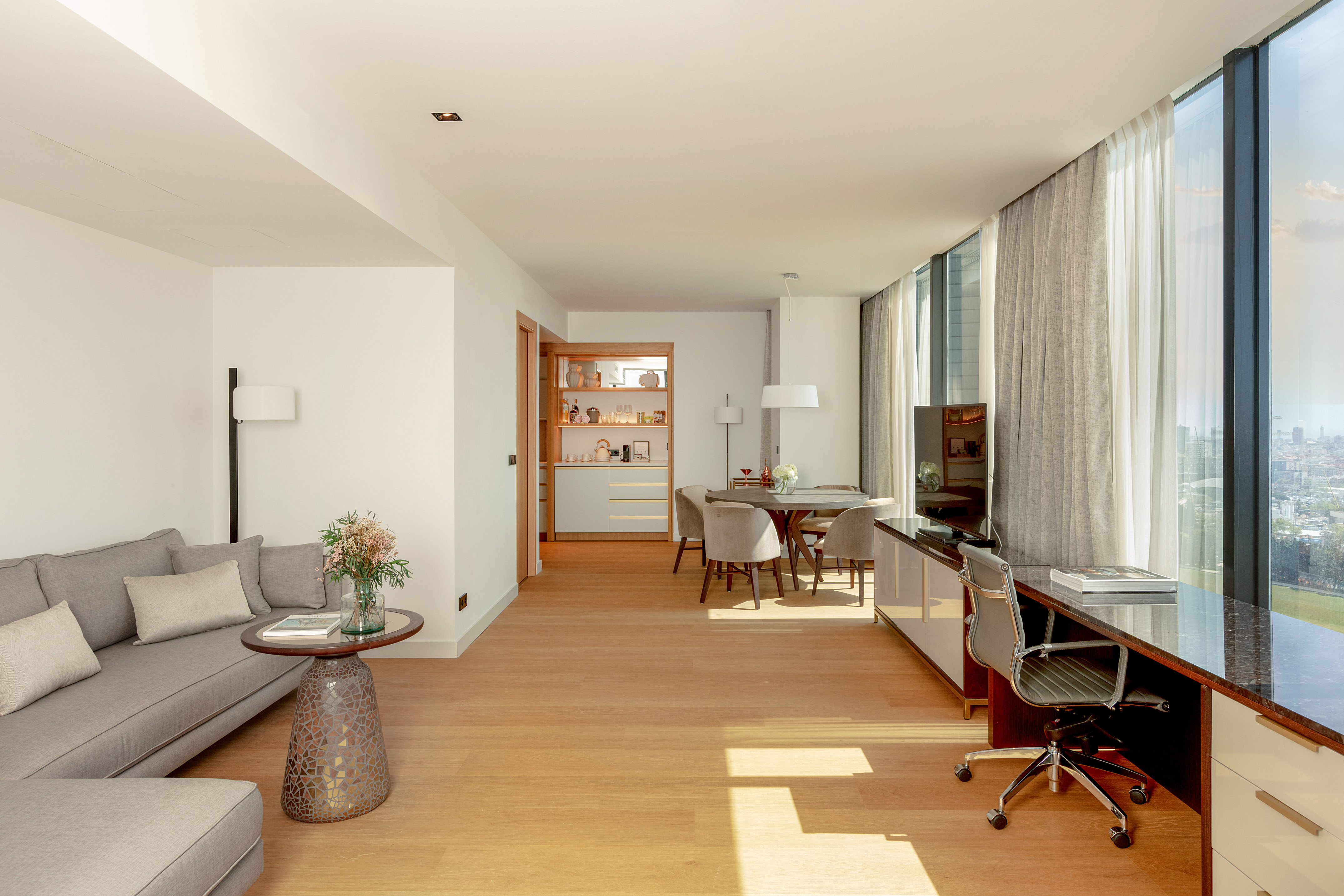 Modern living suite with sofa, dining, desk, hardwood floor, and city view windows.