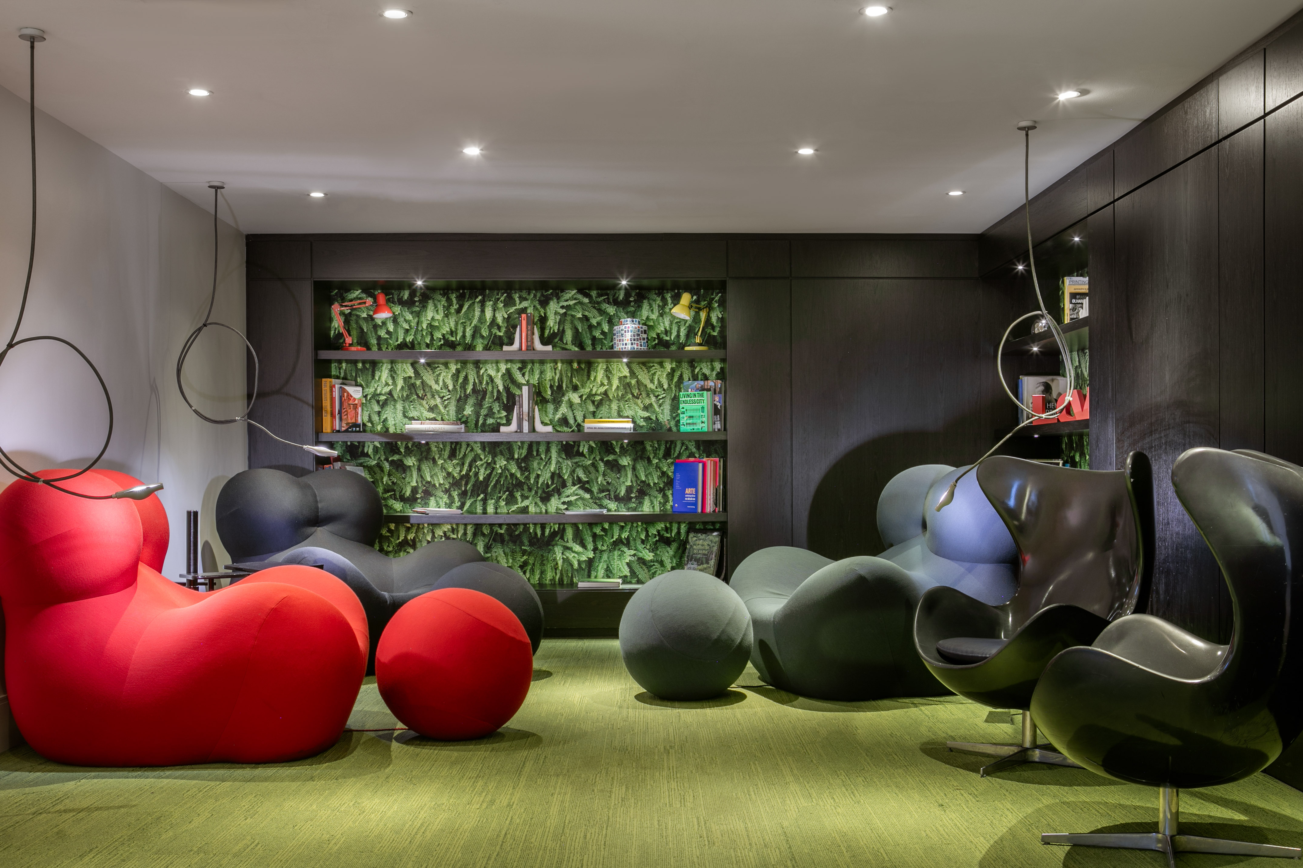 Modern lounge with colorful seating, green carpet, dark wood, and botanical wall shelves.