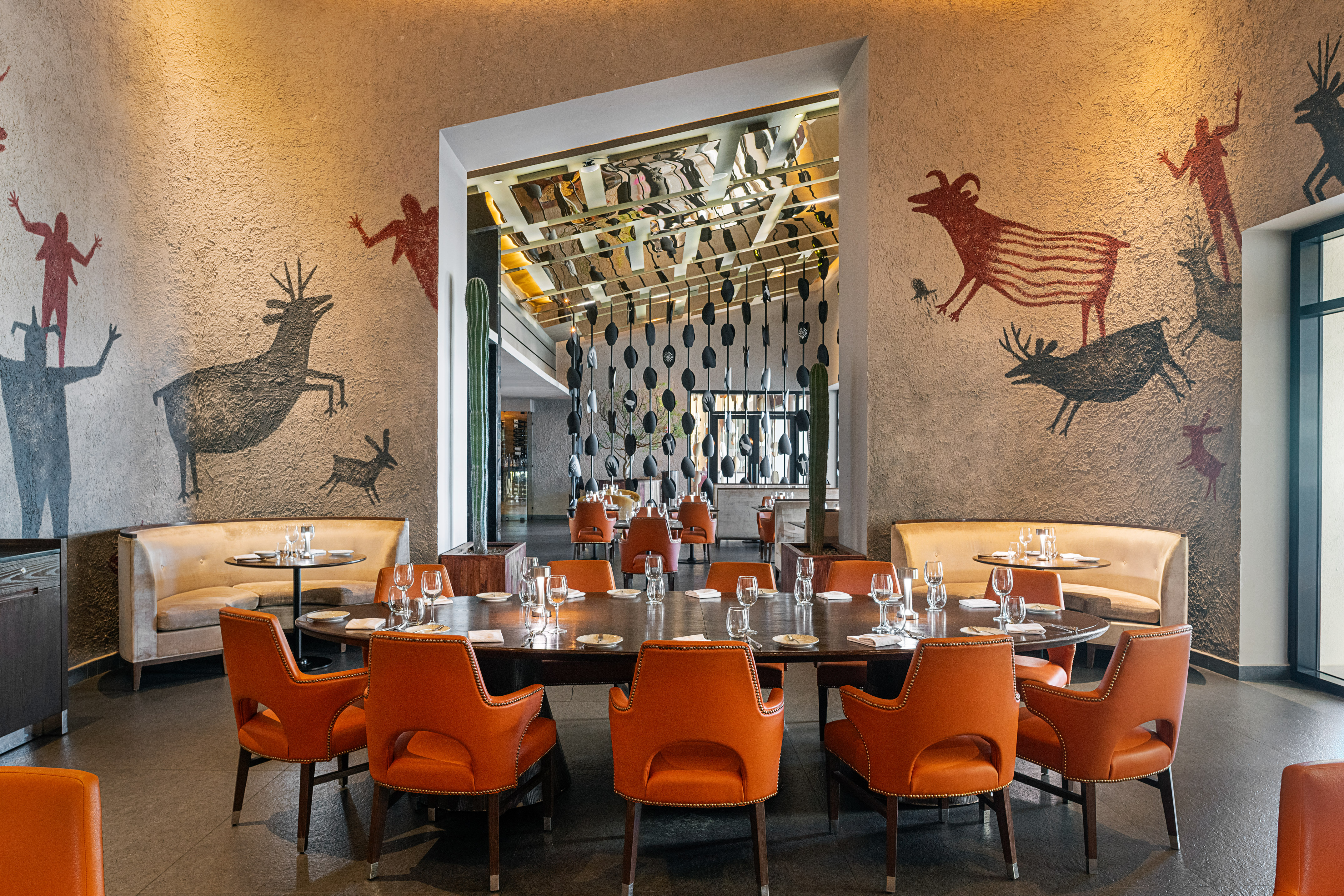 Restaurant interior with cave art murals, orange chairs, and dining tables.