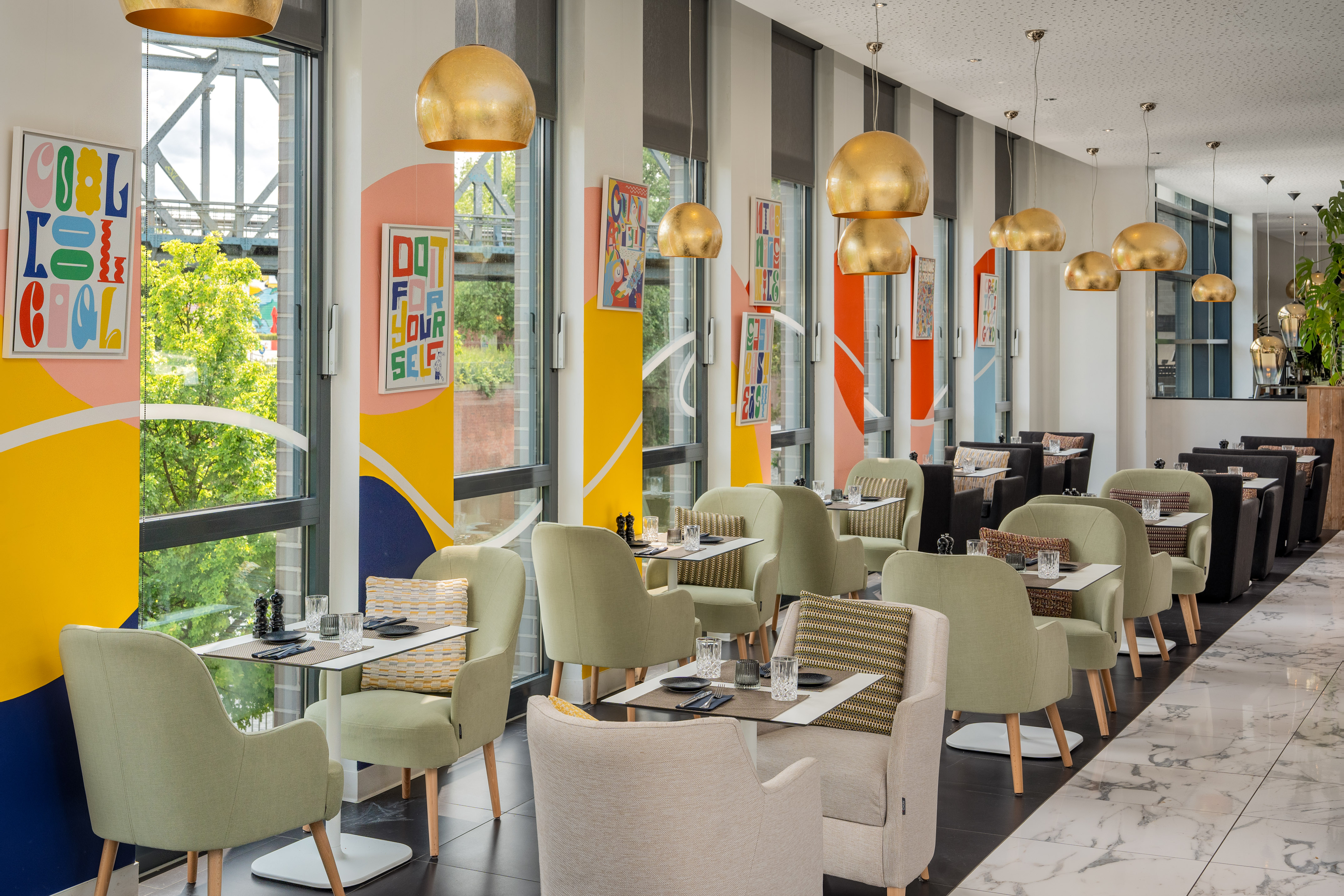 Modern restaurant with vibrant decor, green armchairs, and gold pendant lights.