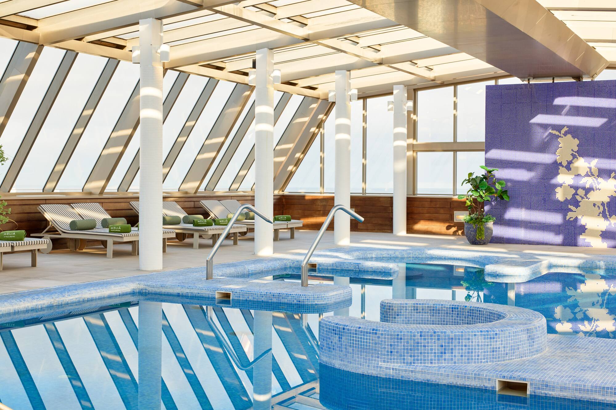 Modern indoor pool with hot tub, blue tiles, lounge chairs, and glass roof.