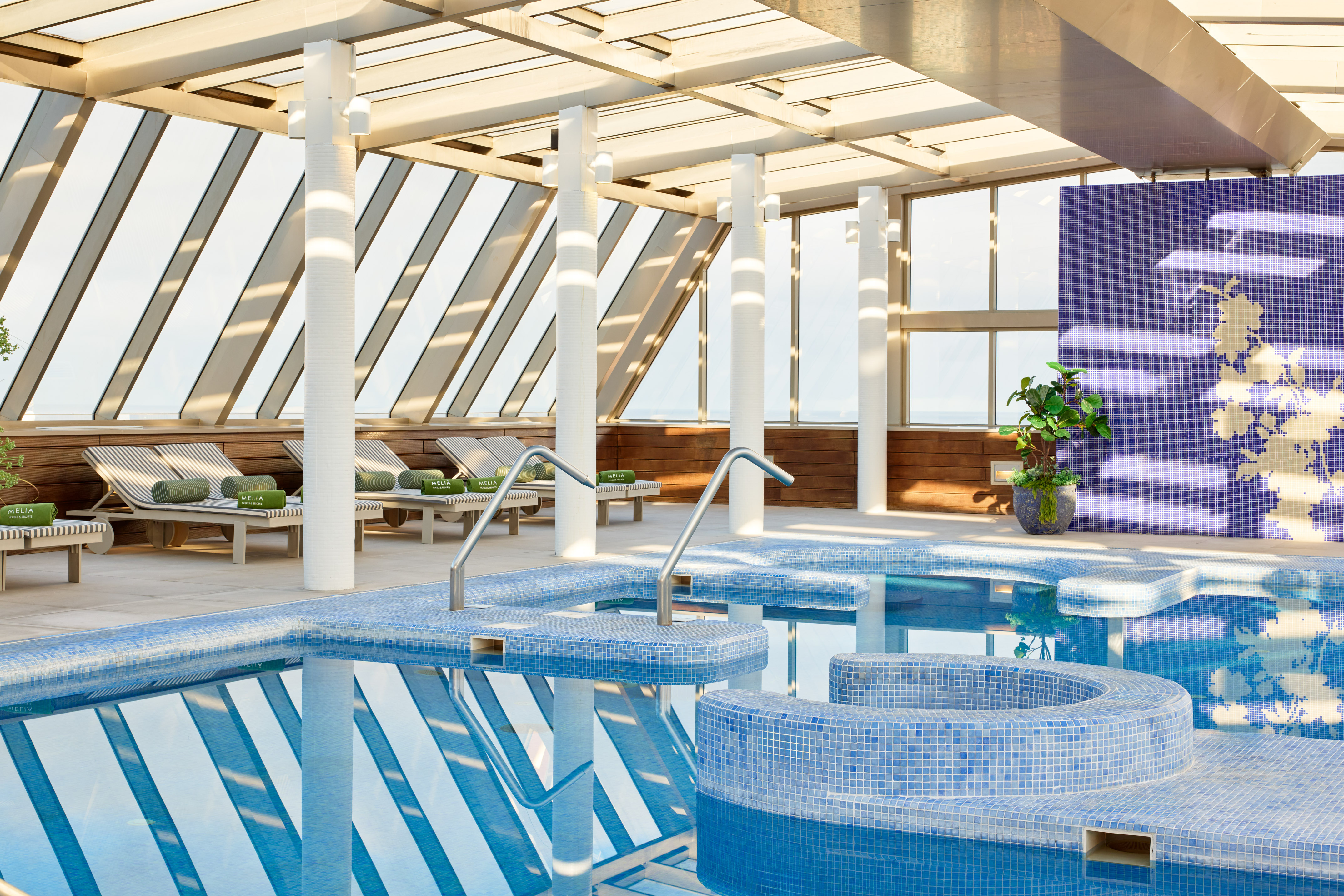 Modern indoor pool with hot tub, blue tiles, lounge chairs, and glass roof.