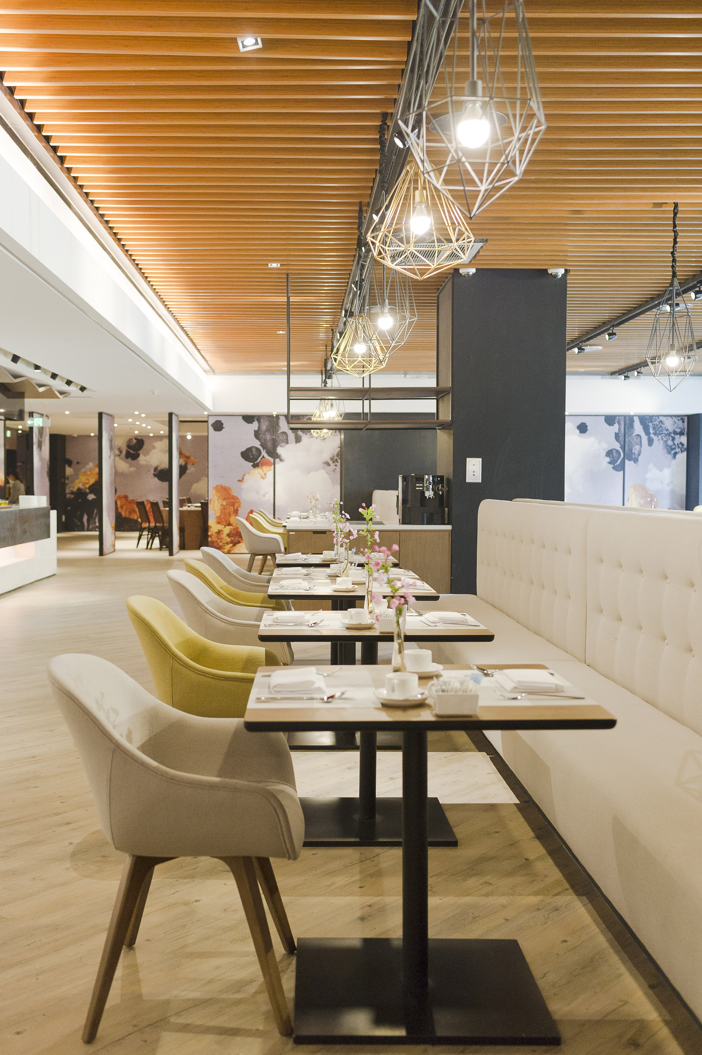 Modern restaurant dining area with set tables, cream/yellow chairs, wooden ceiling.