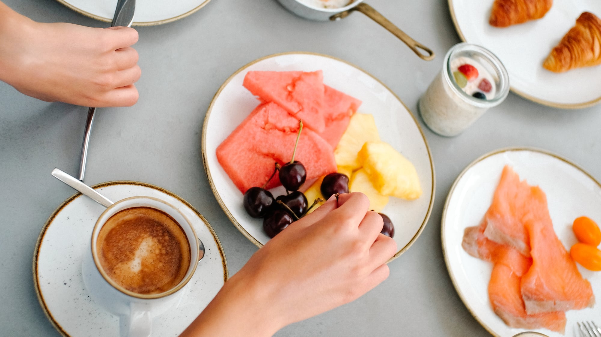 Hands reaching for a varied breakfast spread: coffee, eggs, fruit, salmon, croissants.