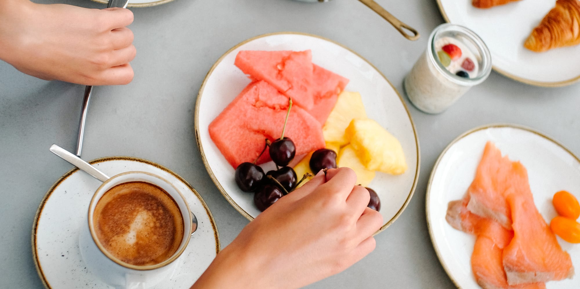 Hands reaching for a varied breakfast spread: coffee, eggs, fruit, salmon, croissants.