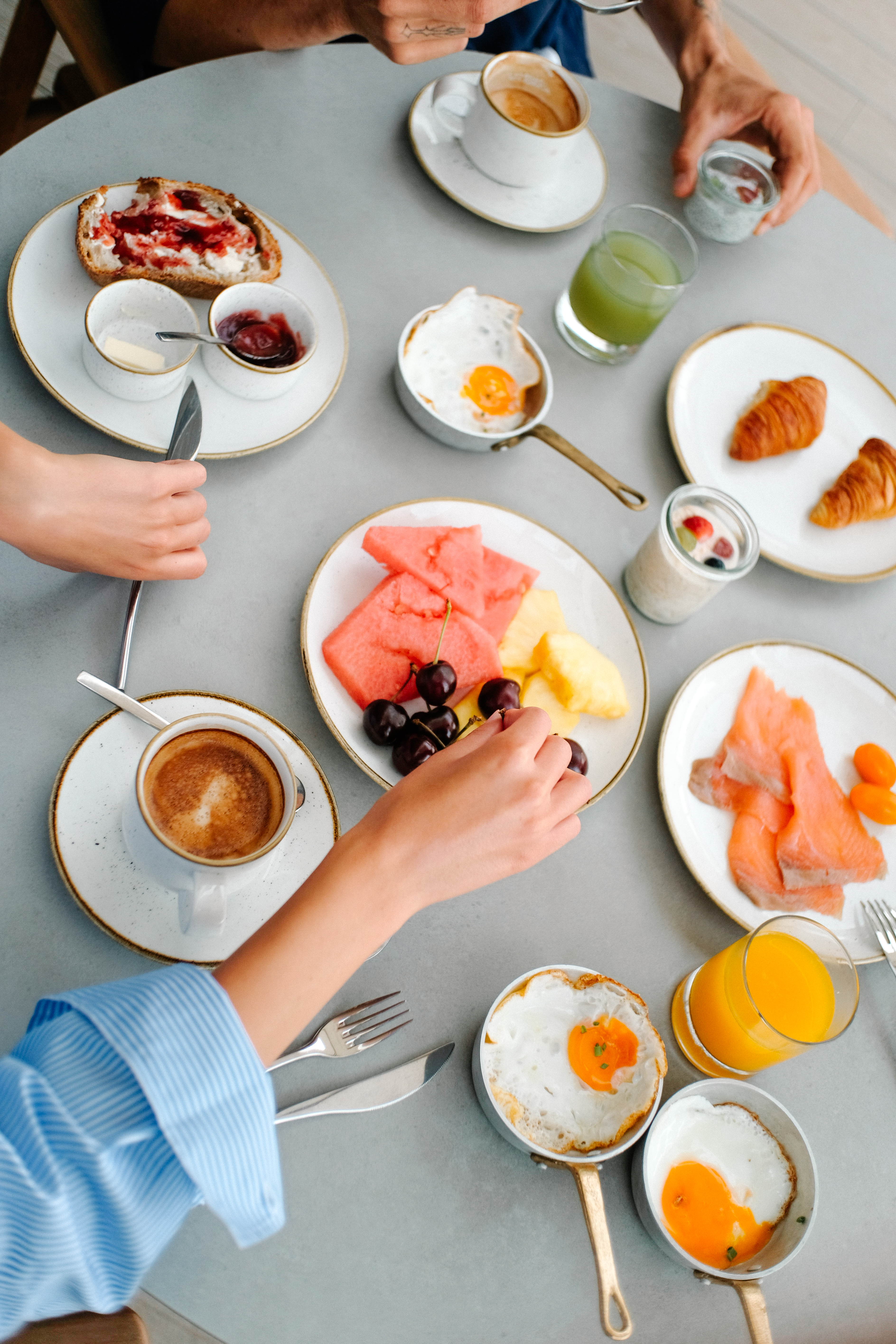 Hands reaching for a varied breakfast spread: coffee, eggs, fruit, salmon, croissants.