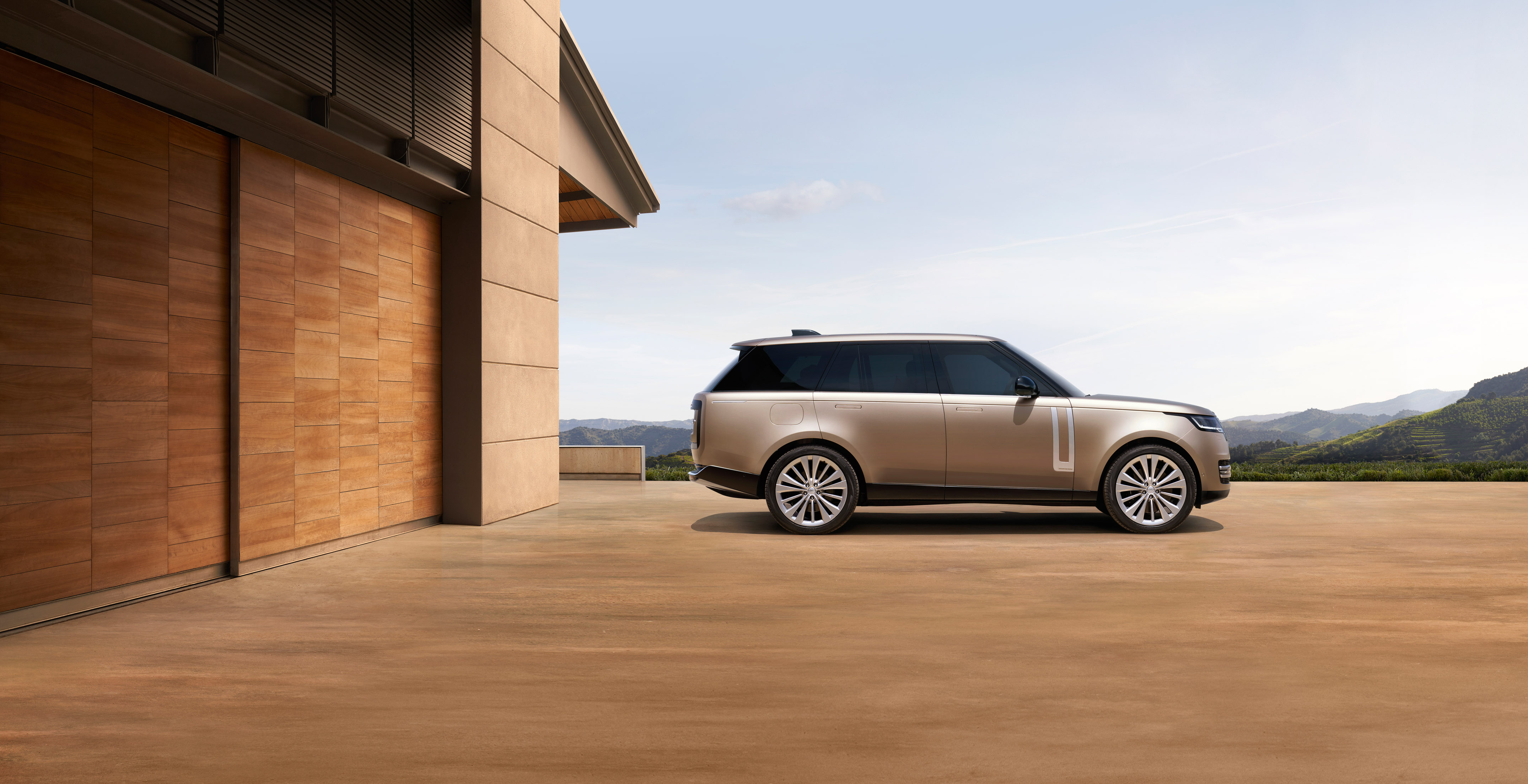 Champagne Range Rover SUV at modern building, mountain backdrop.