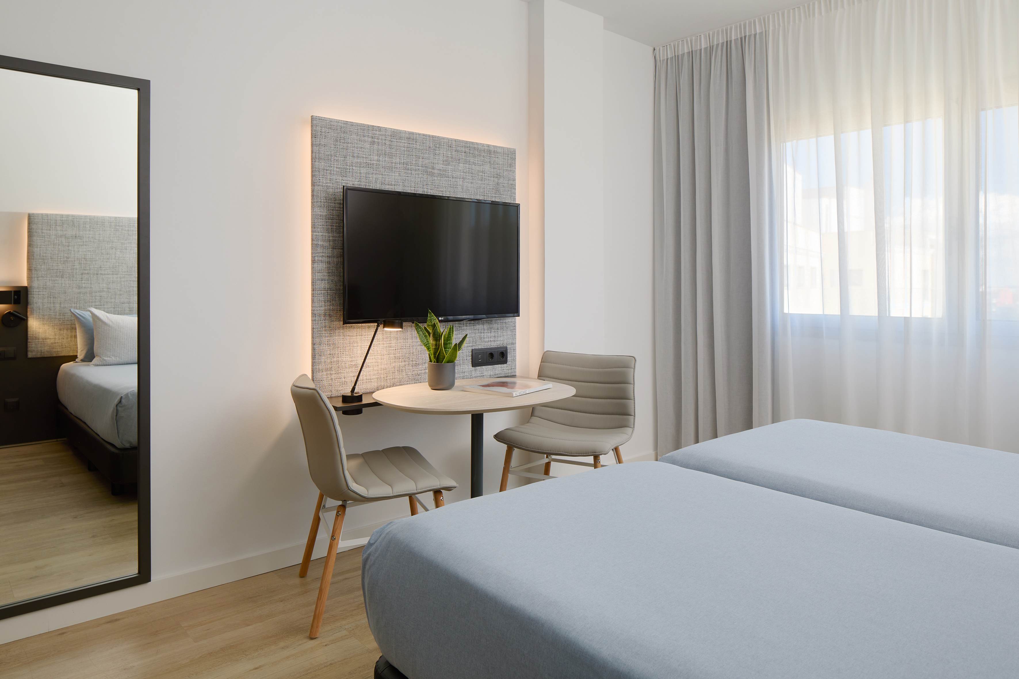Modern twin hotel room with two beds, wall-mounted TV, and small seating area.