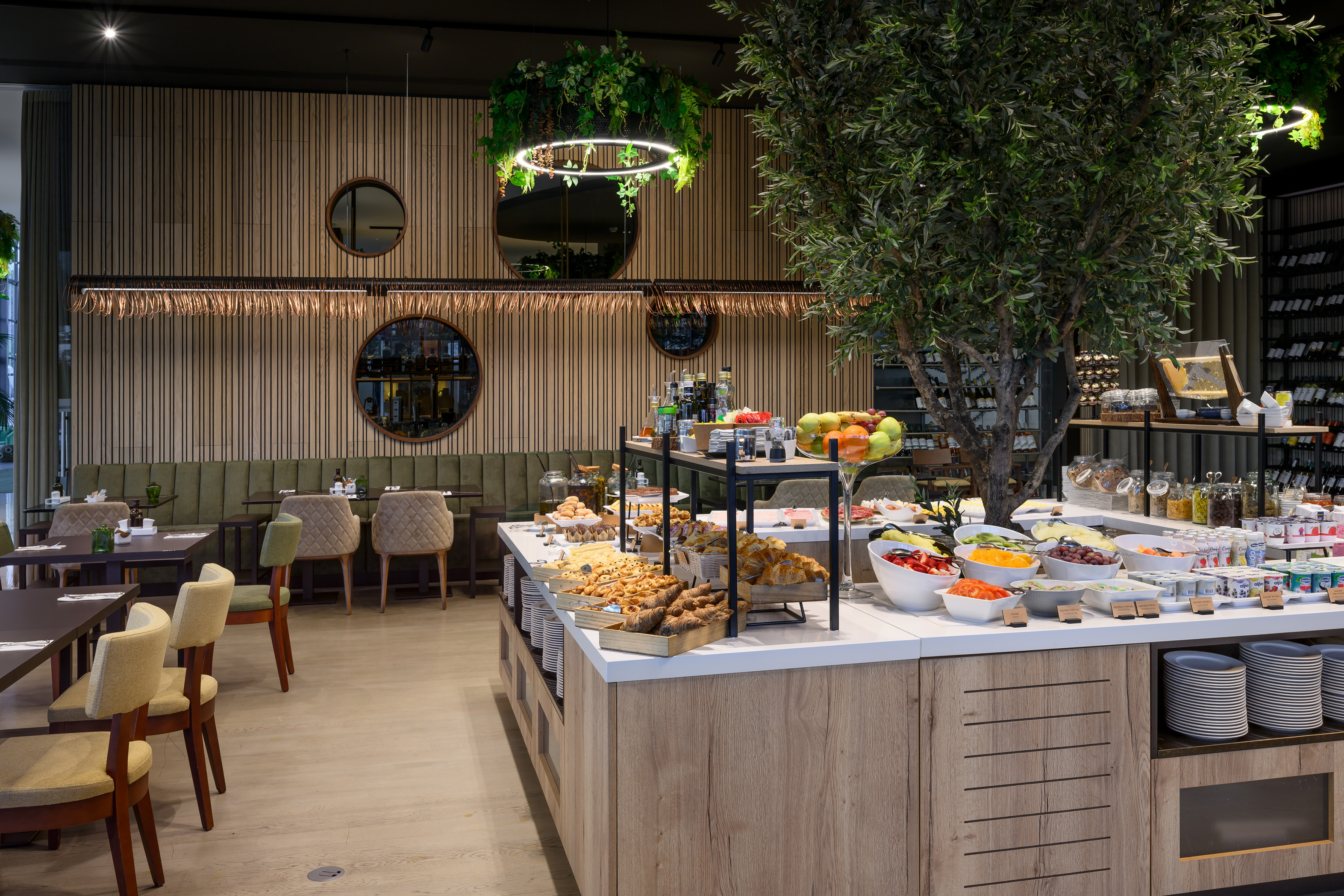 Modern restaurant breakfast buffet with fresh food, olive tree, and stylish decor.