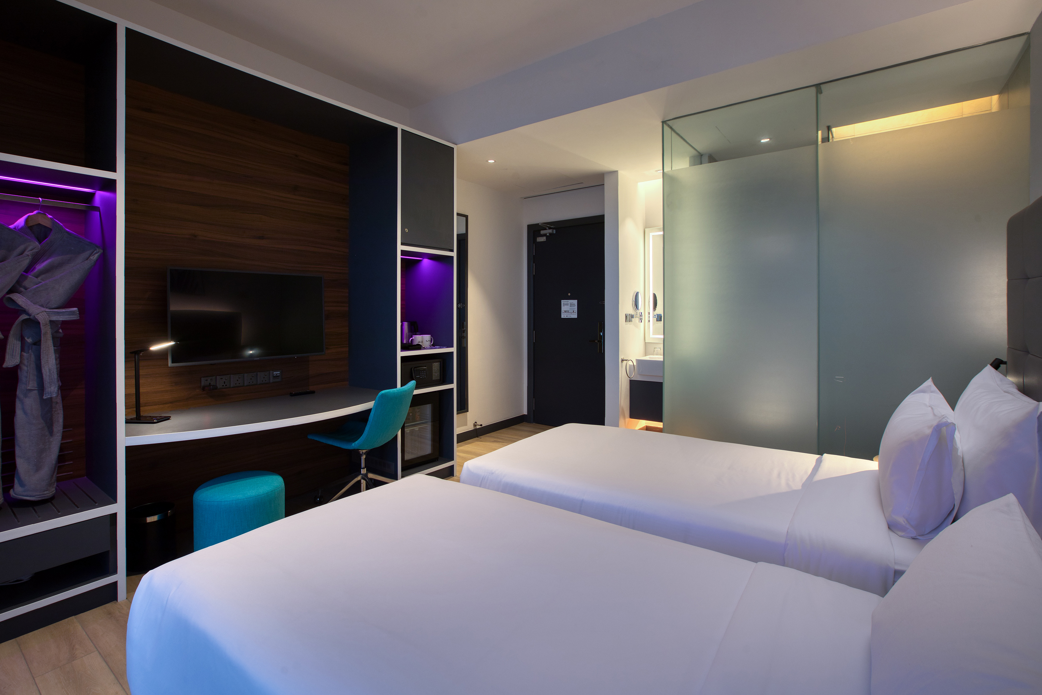 Stylish hotel room with twin beds, desk, TV, and frosted glass bathroom partition.