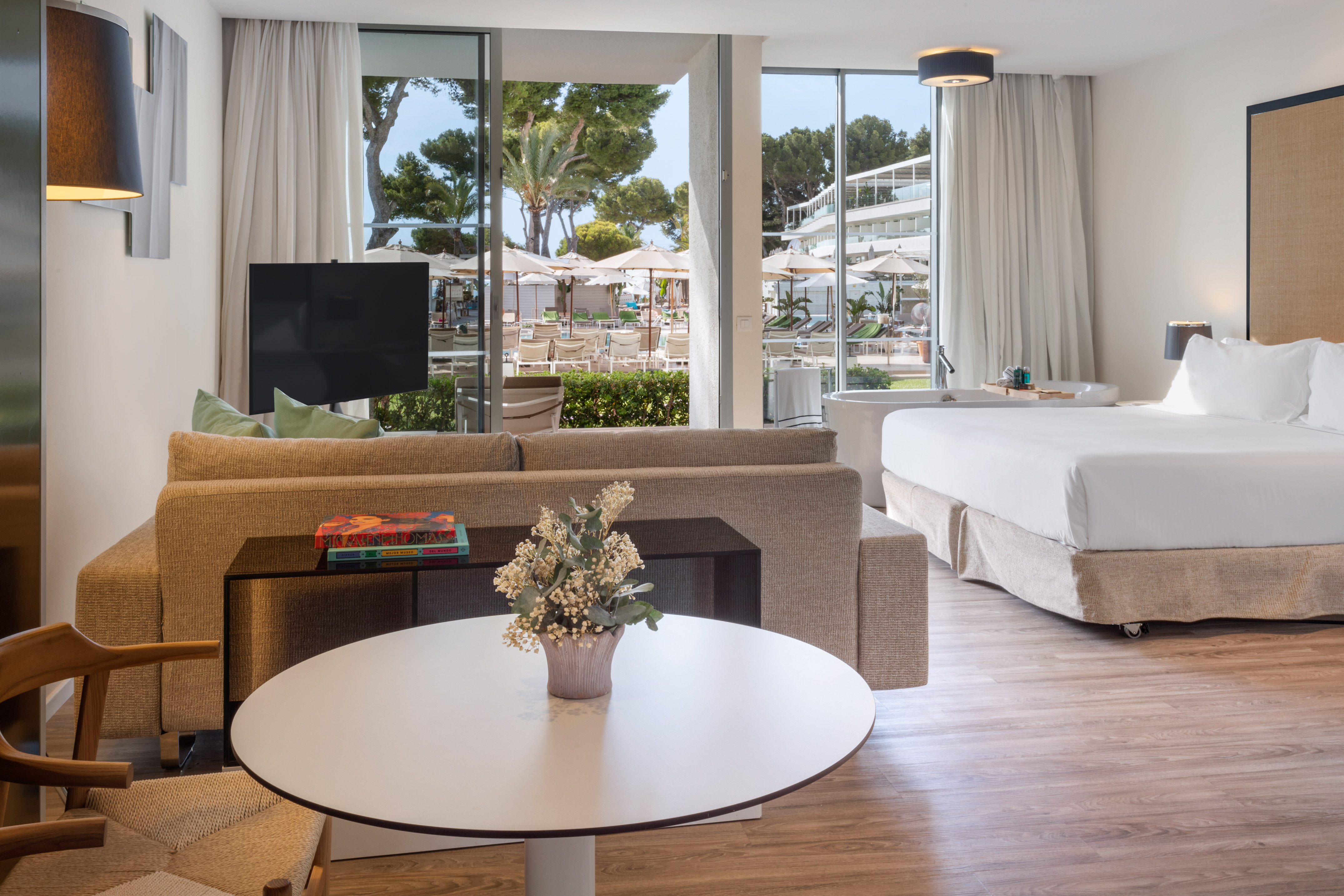 Hotel suite with bedroom, living area, bathtub, and open doors to a resort pool terrace.