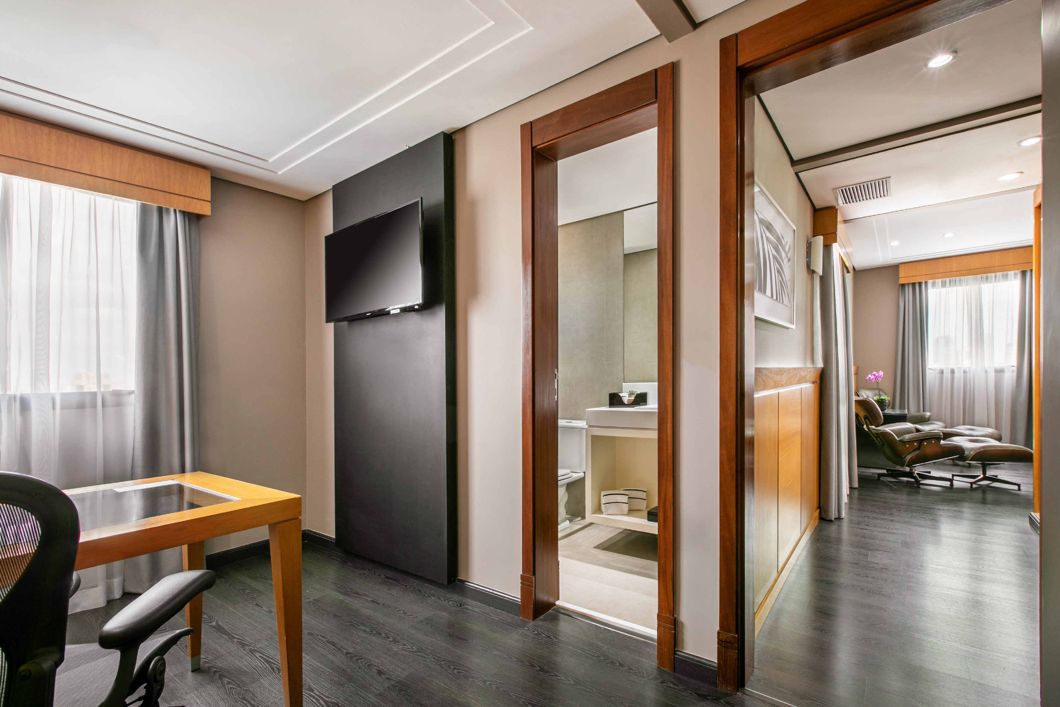 Modern hotel suite with desk, TV, bathroom, and lounge view. Dark wood floor.