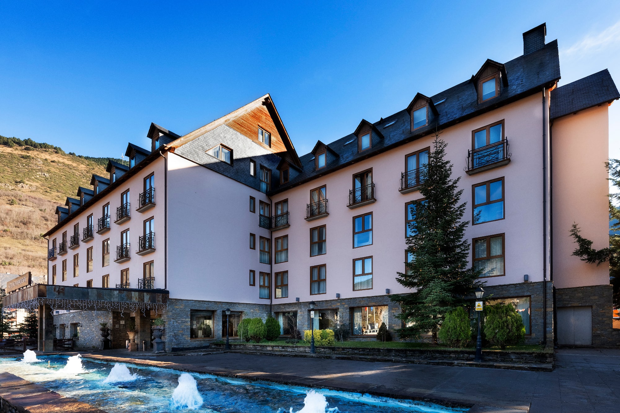 Mountain resort hotel with pink facade, slate roof, balconies, and fountain.