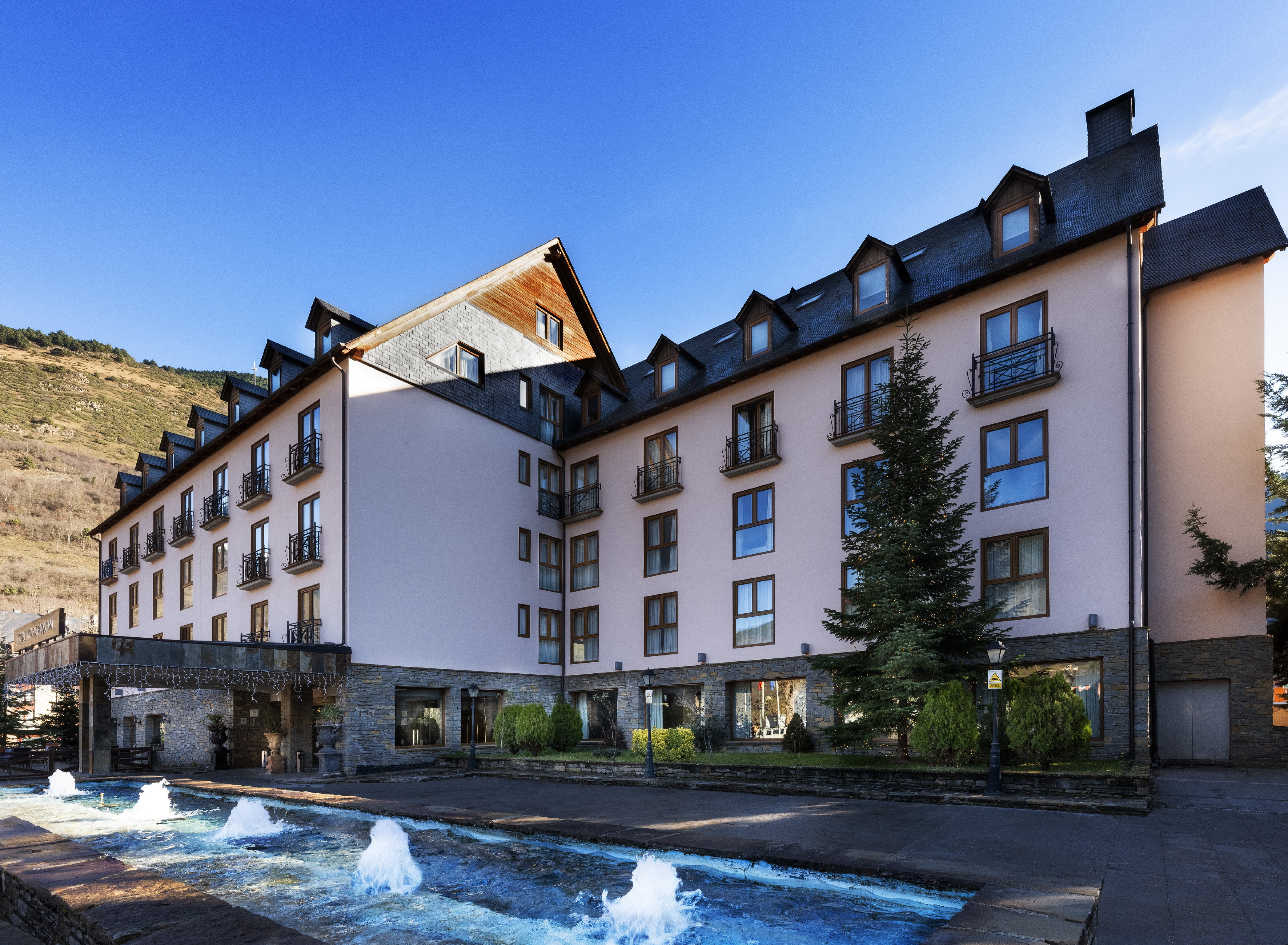 Mountain resort hotel with pink facade, slate roof, balconies, and fountain.