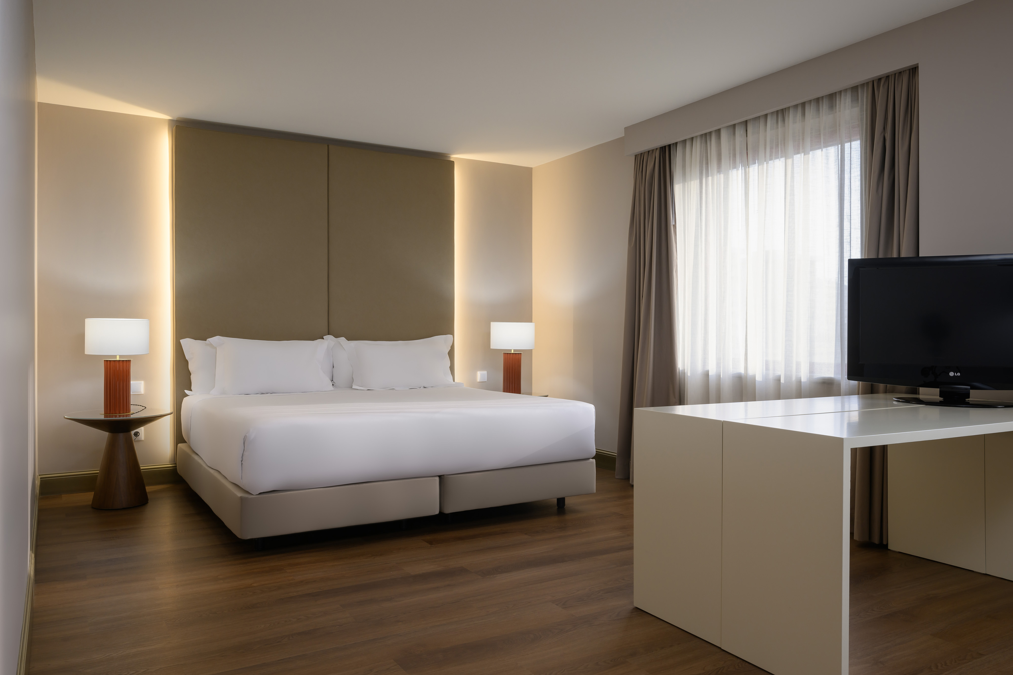 Modern hotel bedroom with large bed, warm backlighting, wood floor, TV, window.