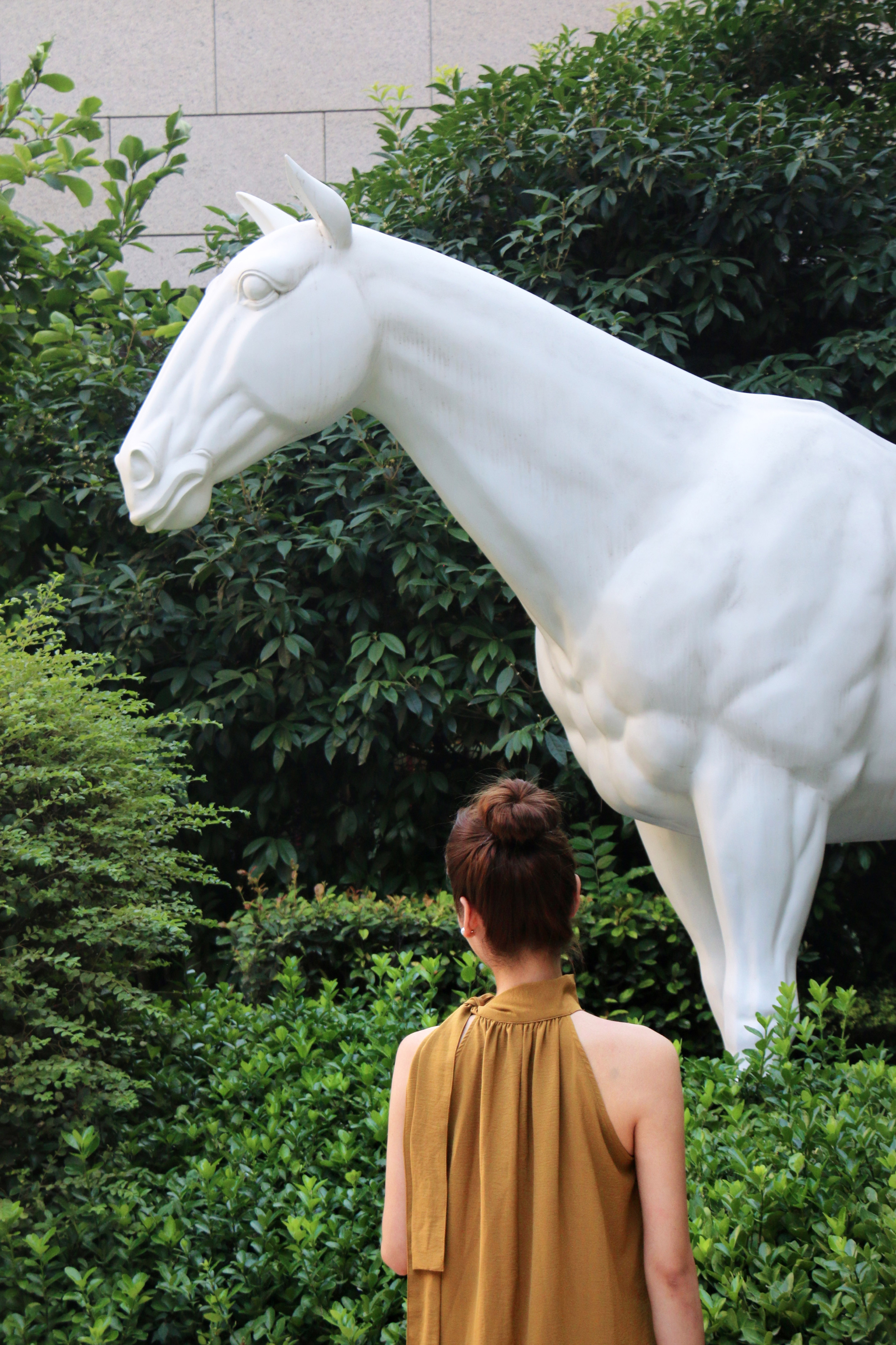 Woman admiring a large white horse sculpture surrounded by lush green foliage.