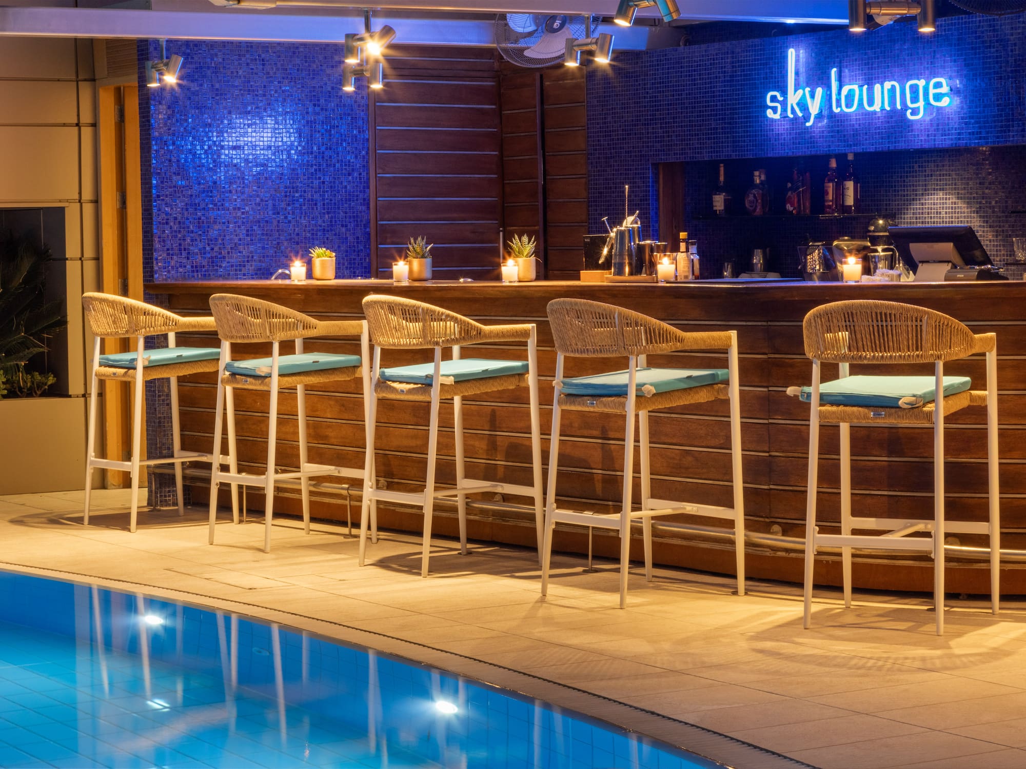 Hotel Sky Lounge bar with neon sign, pool, and wicker stools at night.